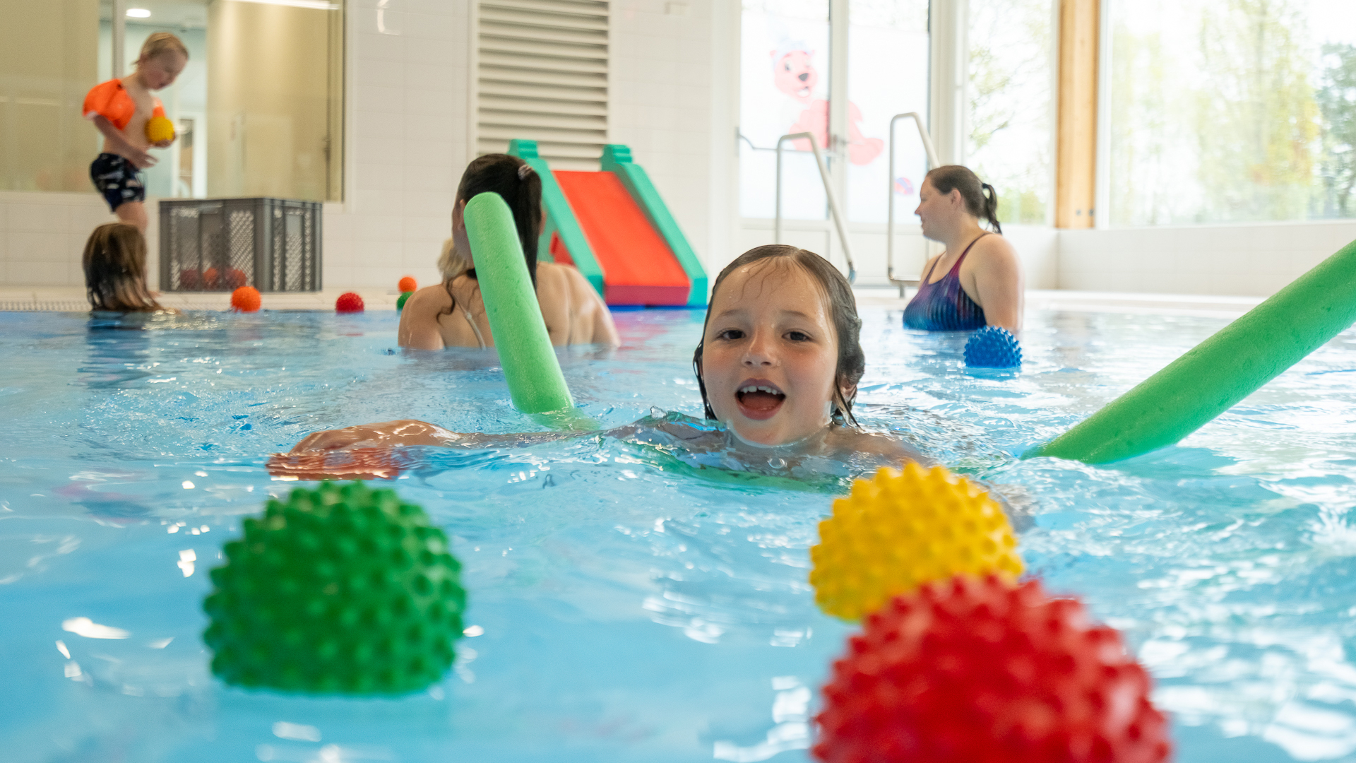 Kinderen spelen in een zwembad met kleurrijke ballen en zwemattributen; op de achtergrond zijn volwassenen en een kleine glijbaan te zien.
