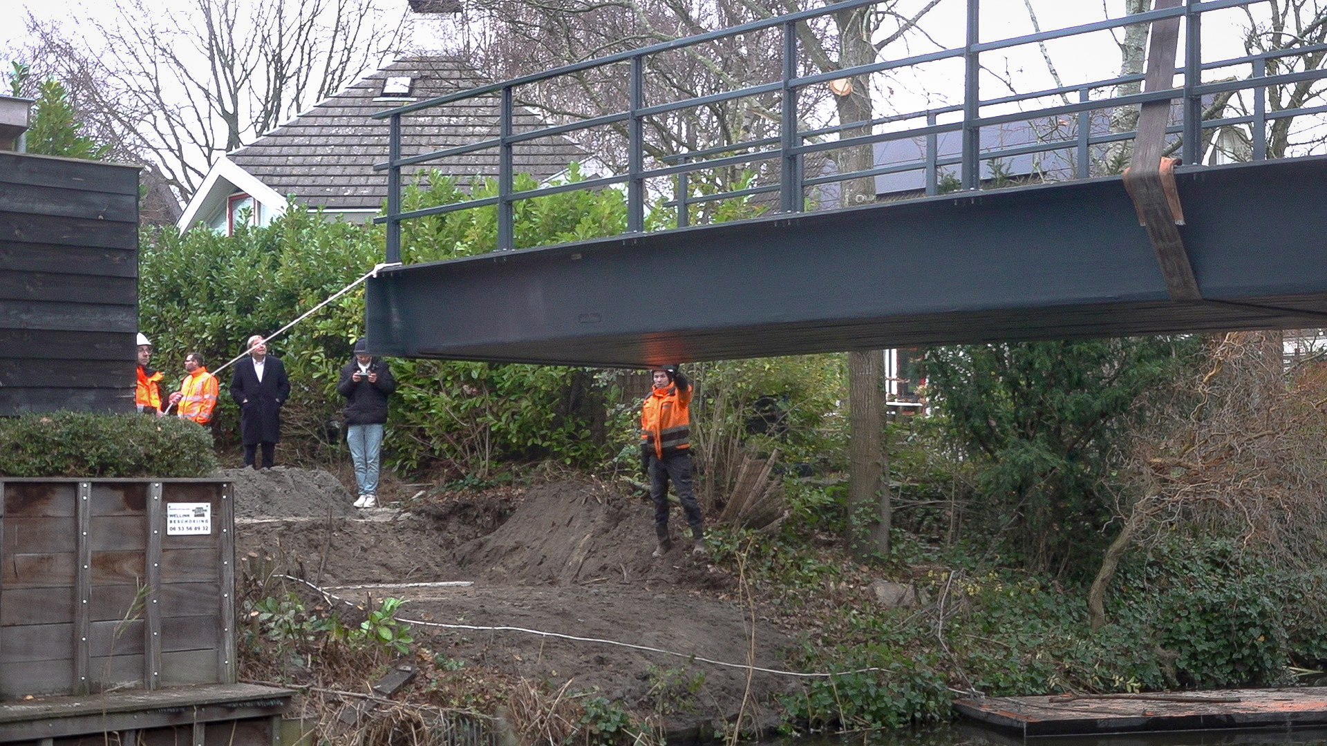 Een metalen brug die wordt geïnstalleerd boven een waterloop, met bouwvakkers in oranje veiligheidsvesten die toezicht houden en toeschouwers in de nabijheid.
