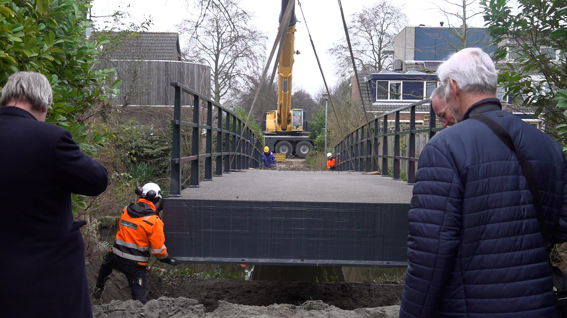 Een brug wordt met behulp van een kraan geplaatst, terwijl meerdere mensen toekijken, waarvan sommigen bouwhelmen en veiligheidskleding dragen.