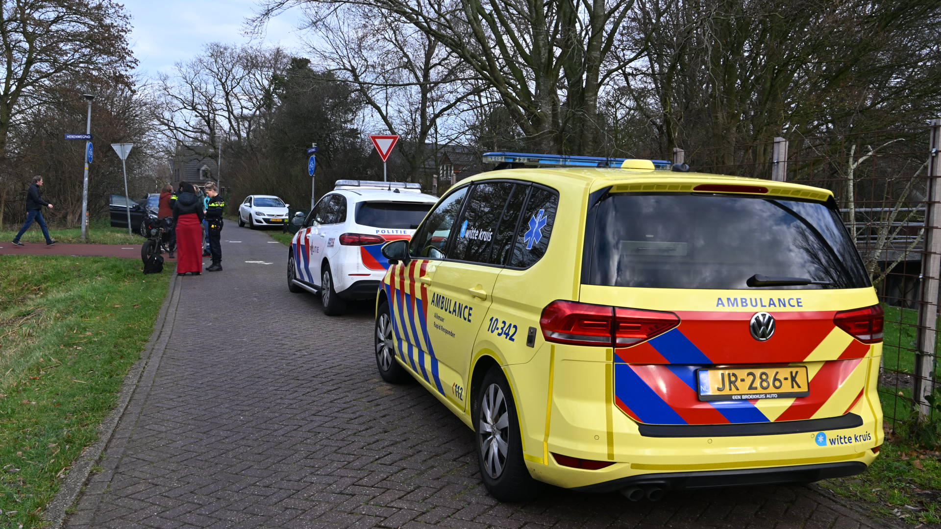 Ambulance en politieauto staan geparkeerd op een smalle weg met mensen, waaronder een agent, die iets bespreken.