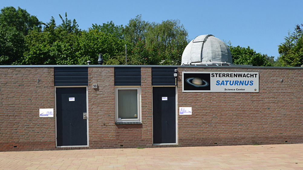 Bakstenen gebouw met de naam "Sterrenwacht Saturnus" op een bord, uitgerust met een koepelvormig observatorium.
