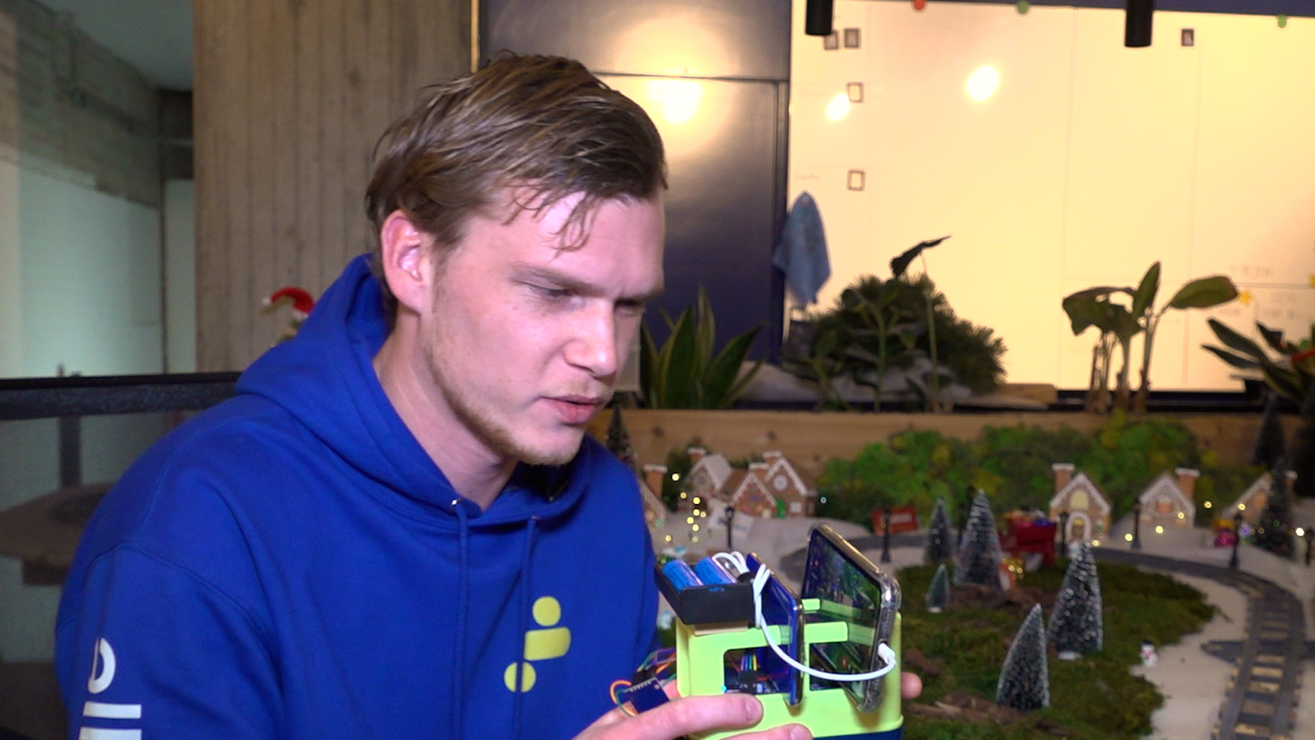 Man in blauwe hoodie onderzoekt een apparaat met batterijen en een scherm, voor een diorama met miniatuurhuizen en bomen.