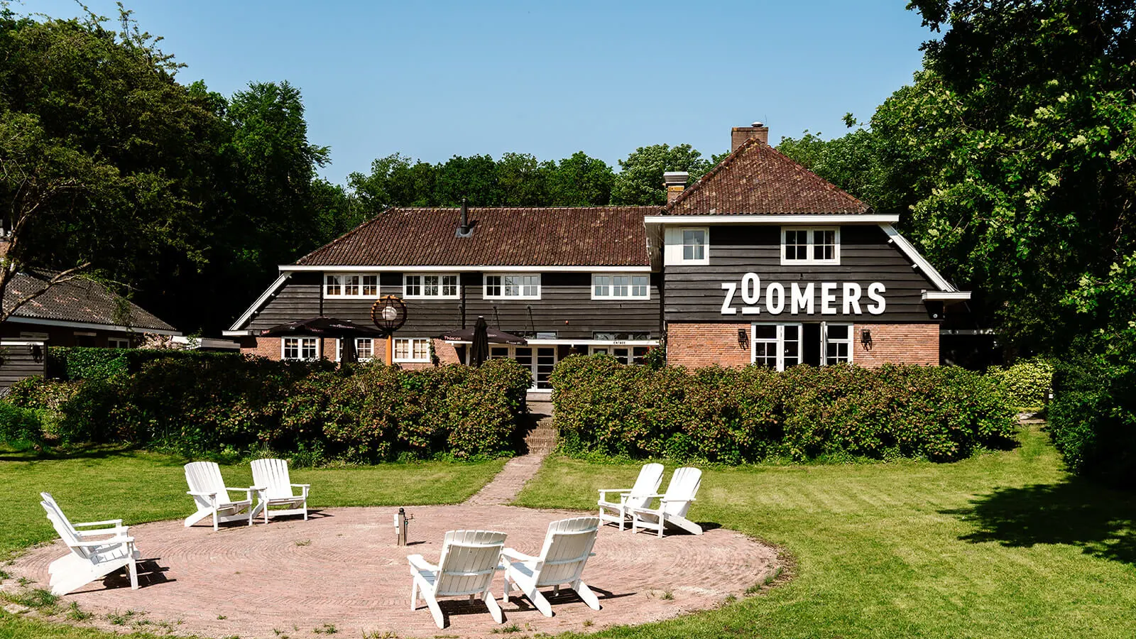 Groot gebouw met houten gevel en grote letters "Zoomers", omgeven door groen met witte stoelen in de tuin.