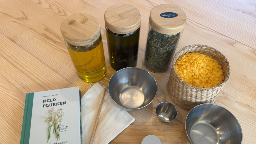 Boodschappen en kookbenodigdheden op een houten tafel, inclusief een boek “Wild Plukken”, potten met olie en gedroogde kruiden, en metalen kommen.