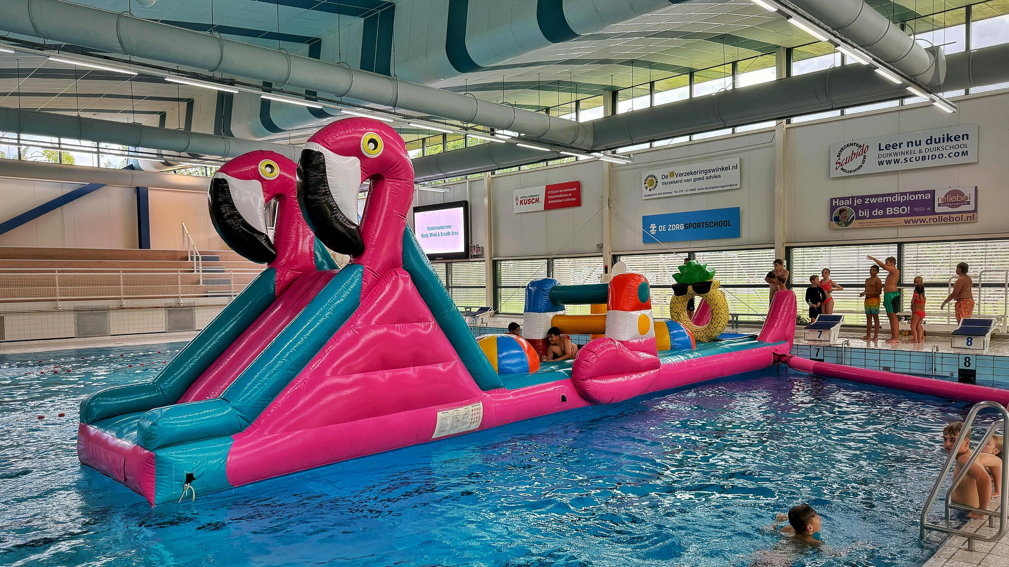 Opblaasbare flamingo-glijbaan in een binnenzwembad met kinderen die klaarstaan op startblokken om erop te spelen.
