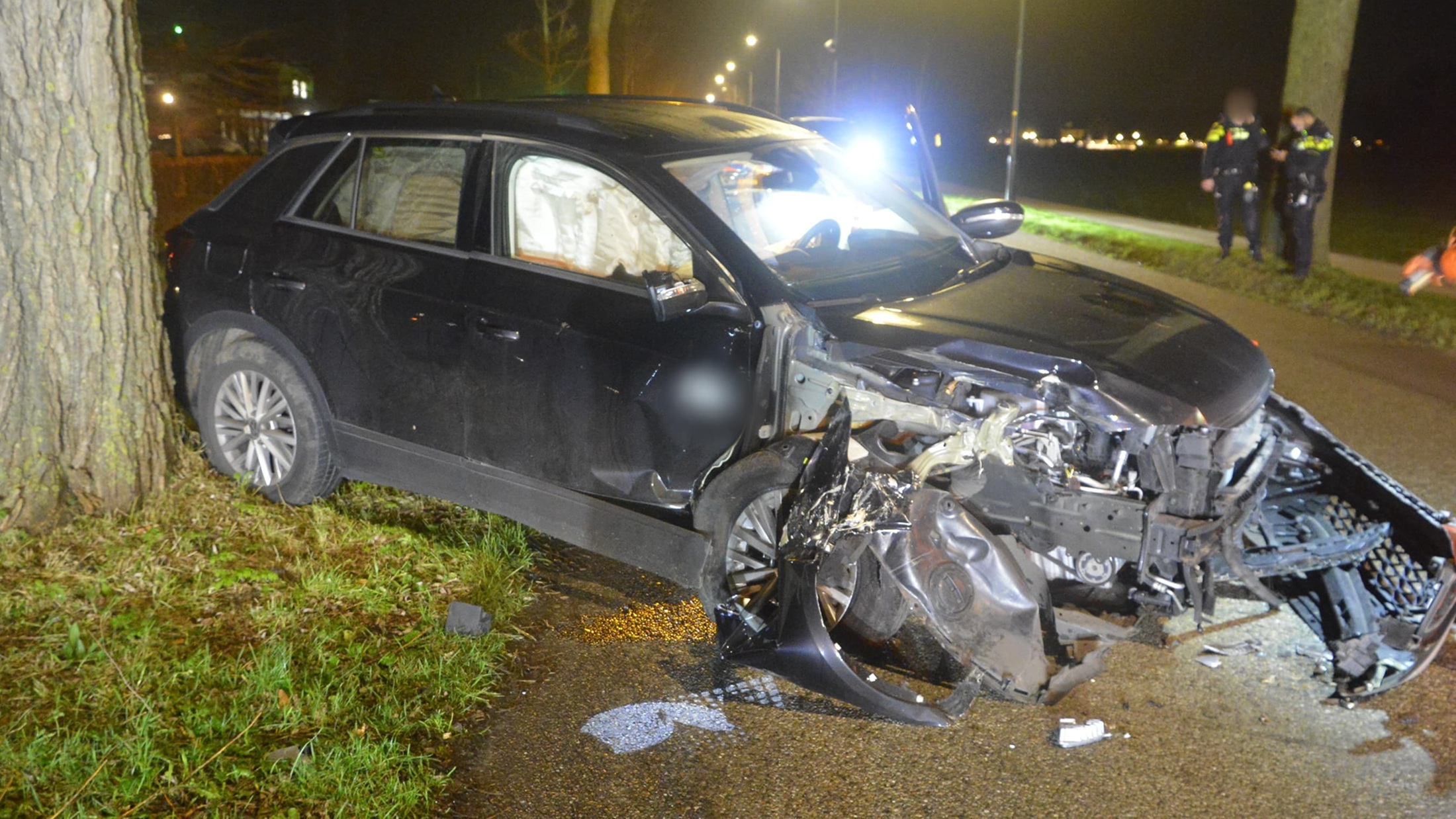 Zwarte auto met zware schade aan de voorkant na botsing tegen een boom; politie en omstanders op de achtergrond.