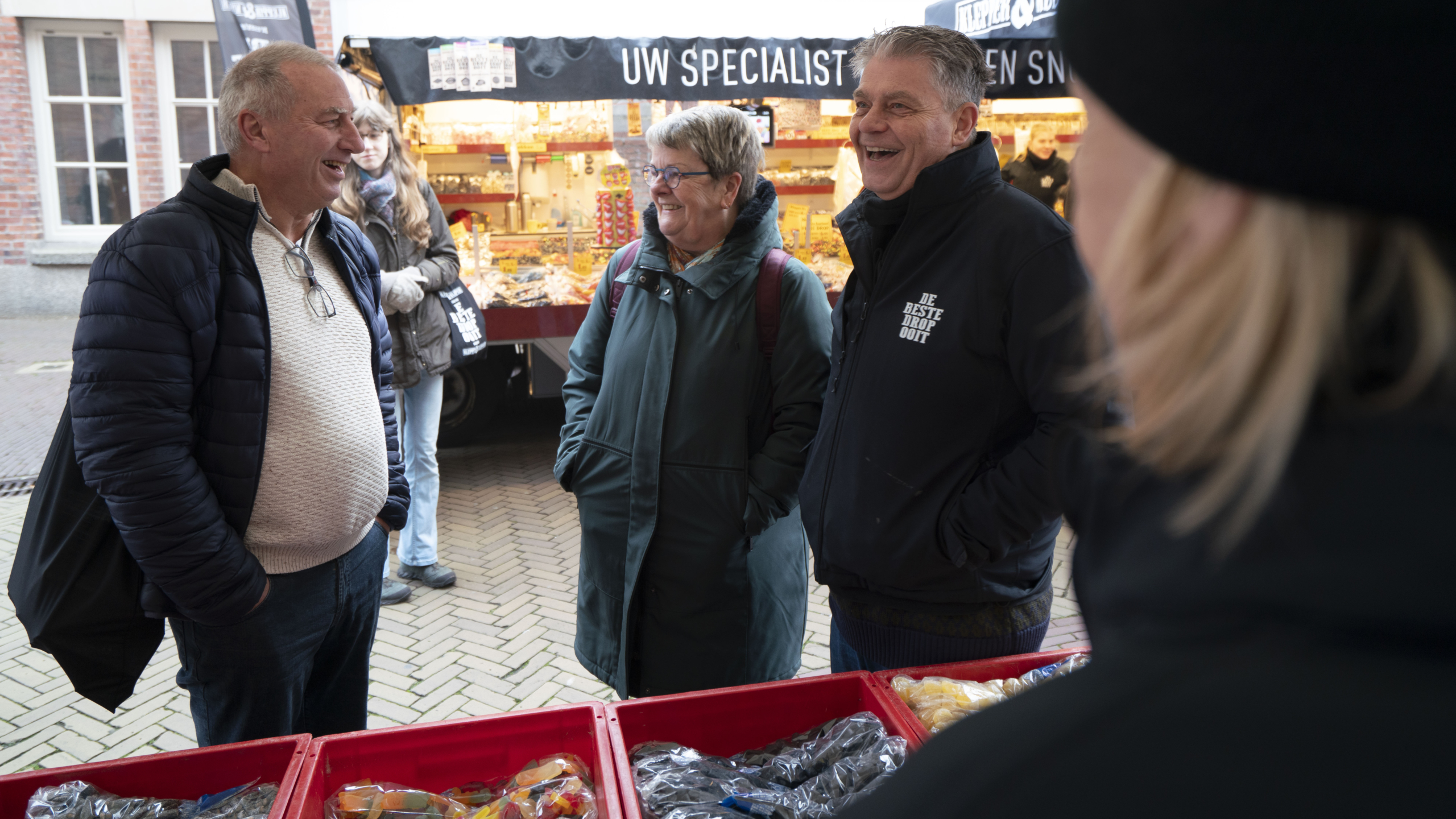 Groep mensen lacht en praat voor een snoepkraam op een markt, met rood gekleurde bakken vol snoep vooraan.