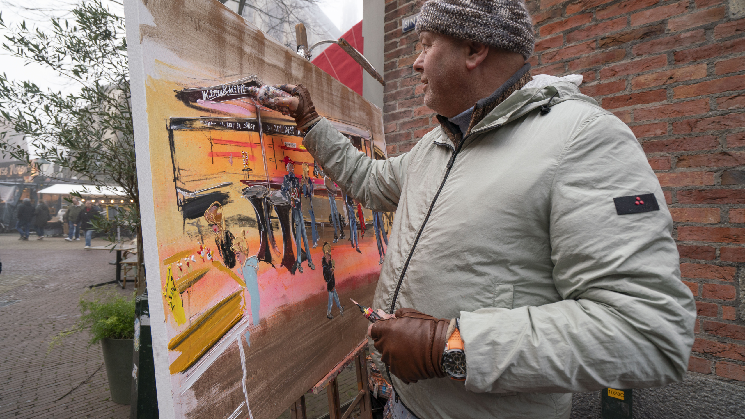 Kunstschilder buiten bezig met een kleurrijk stadsgezicht; draagt een beige jas en wollen muts.