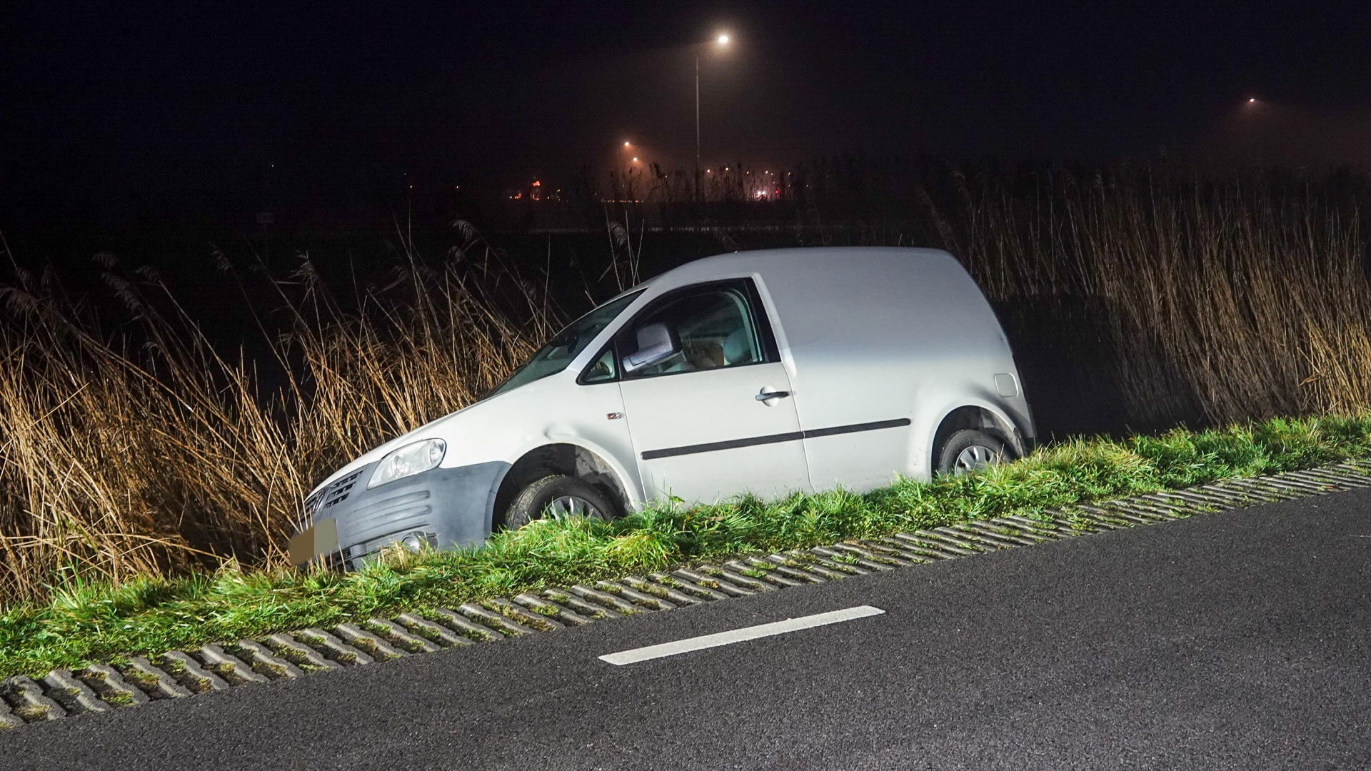 Een witte bestelwagen is van de weg geraakt en ligt half in een greppel naast een donkere weg.