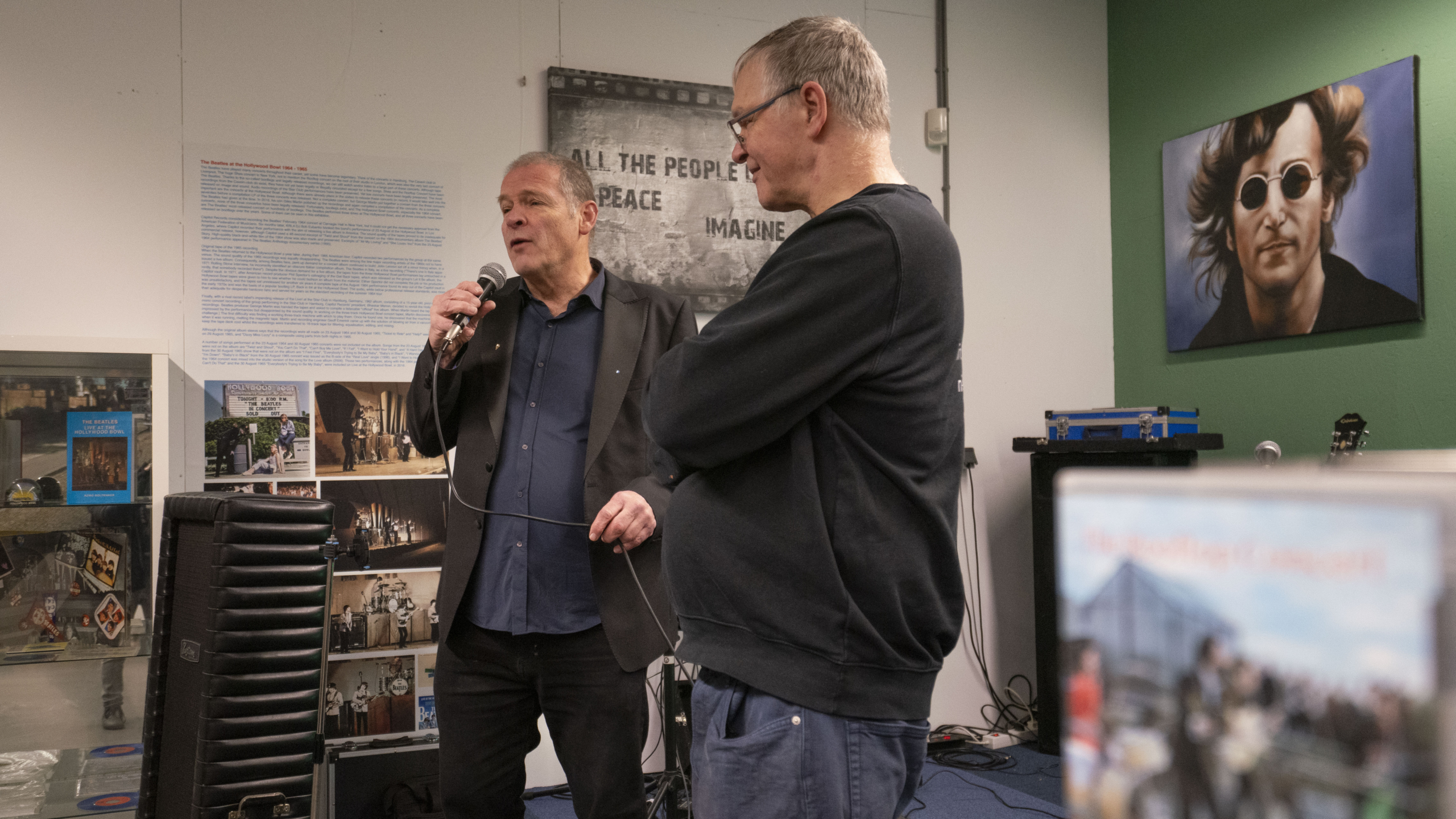Twee mannen praten in een kamer met posters en een schilderij van een man met zonnebril aan de muur.
