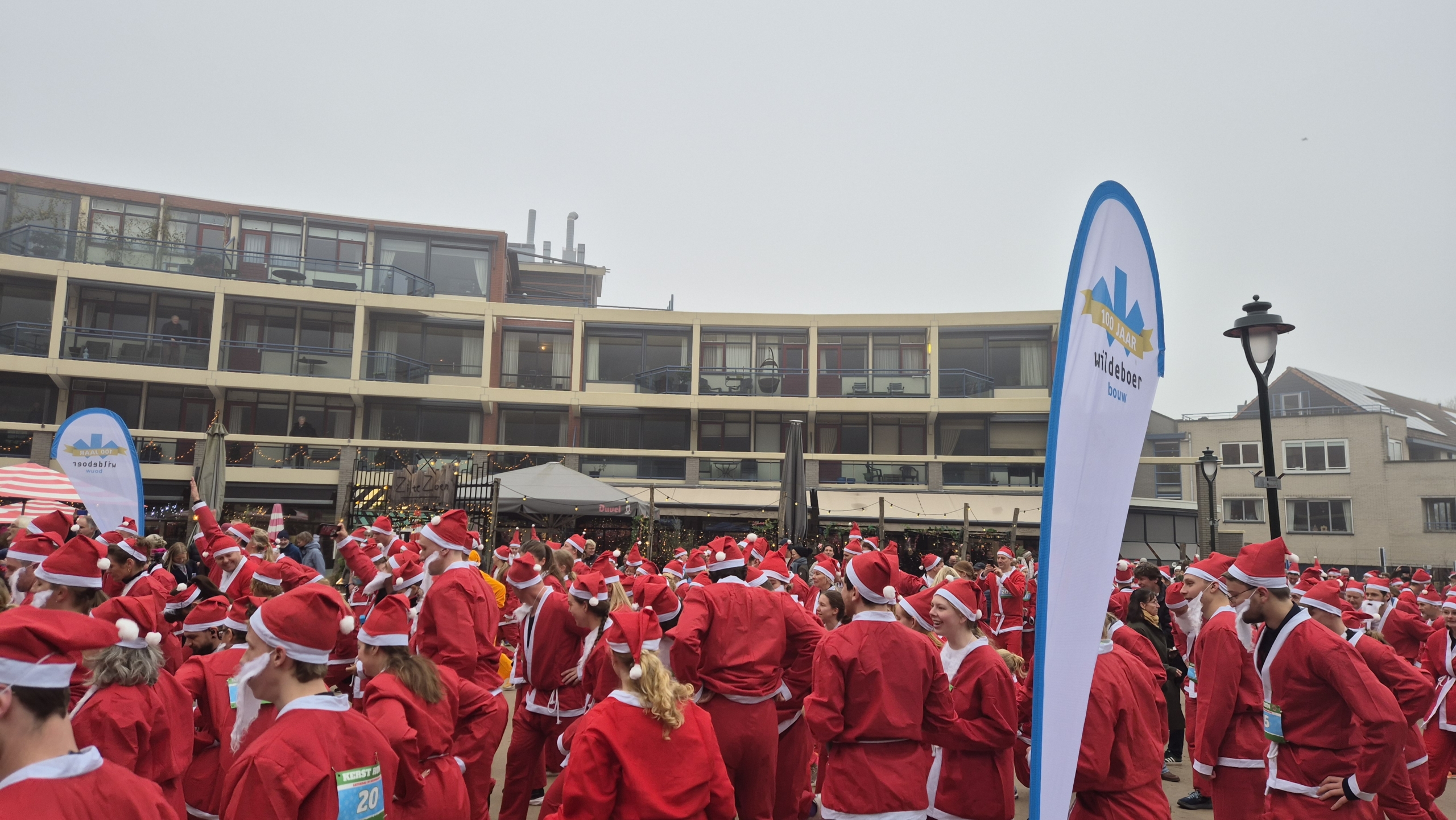 Mensen verkleed als kerstmannen verzamelen zich buiten bij een gebouw met een feestelijke sfeer.