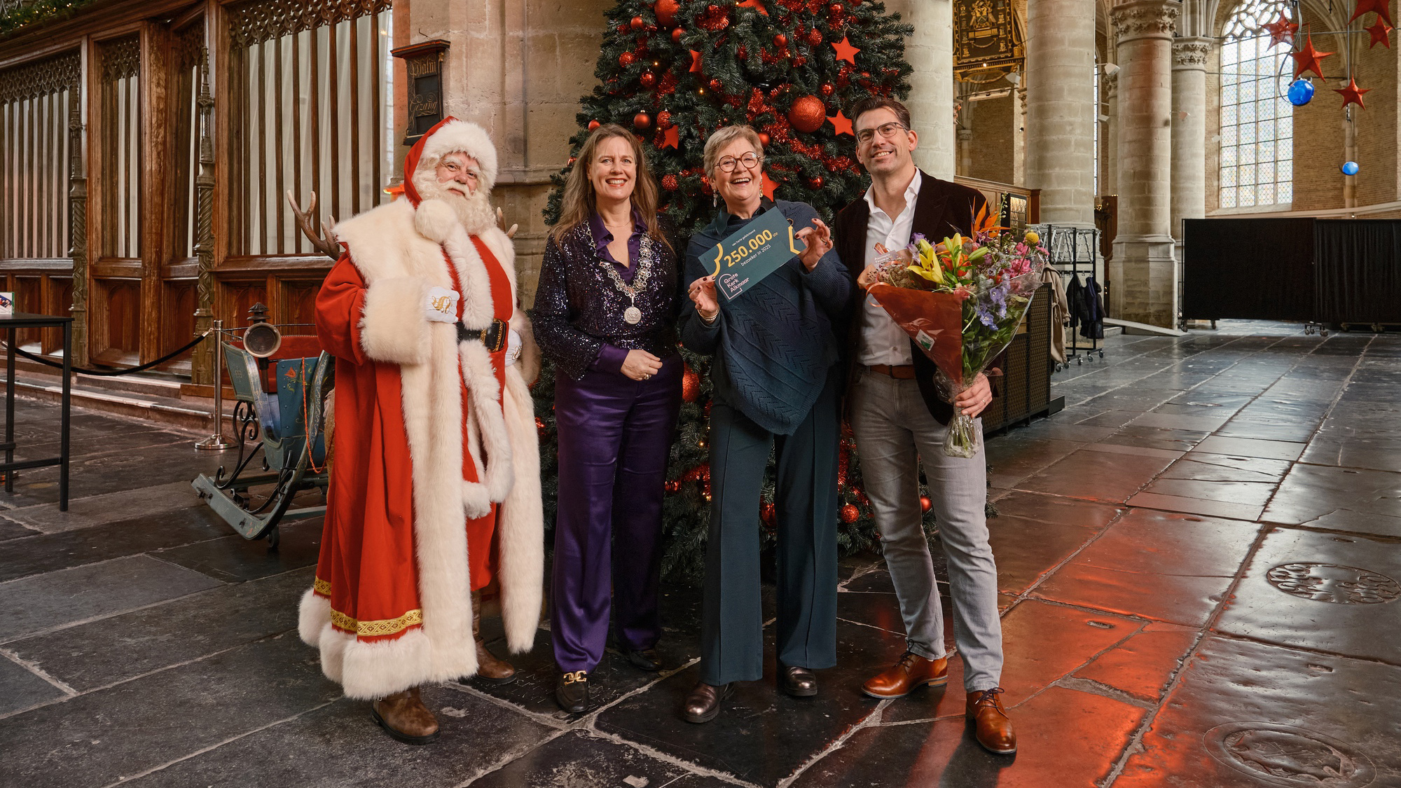 Vier mensen poseren voor een kerstboom in een grote hal, één houdt een cheque vast, een ander een boeket bloemen.