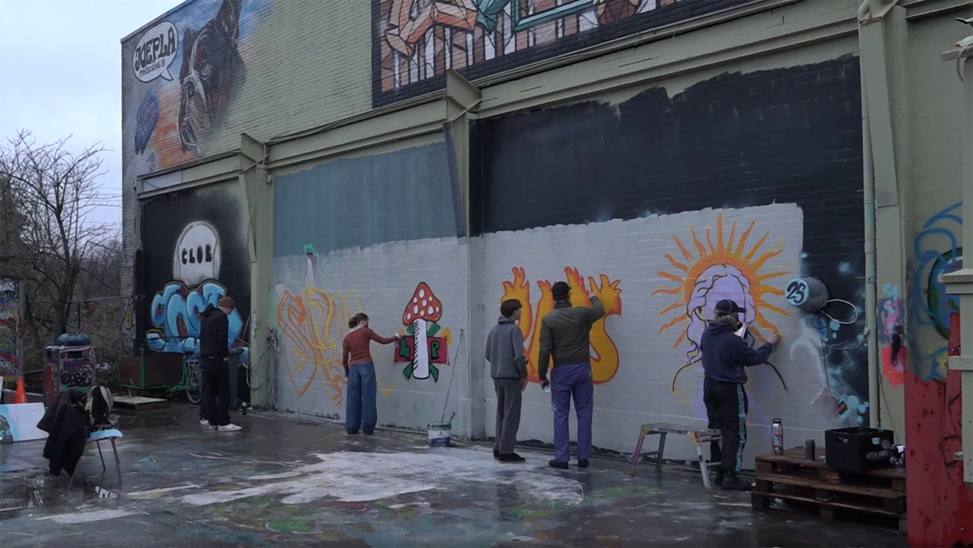 Mensen schilderen samen graffiti op een muur met diverse ontwerpen, waaronder een paddenstoel en een zon.
