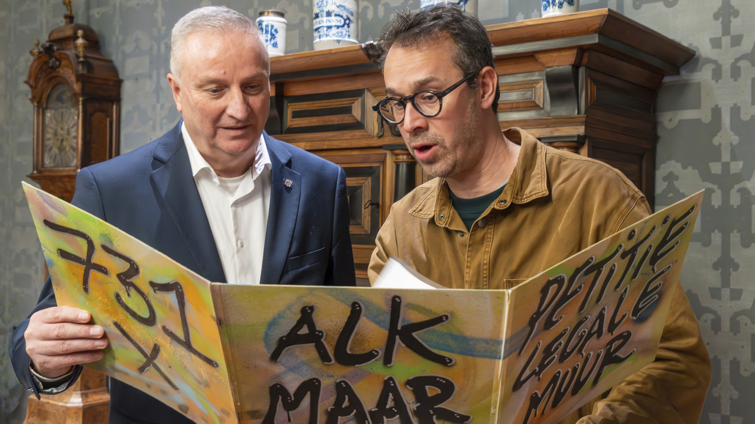 Twee mannen kijken naar een kleurrijk bord met graffiti-tekst in een kamer met antiek meubilair.