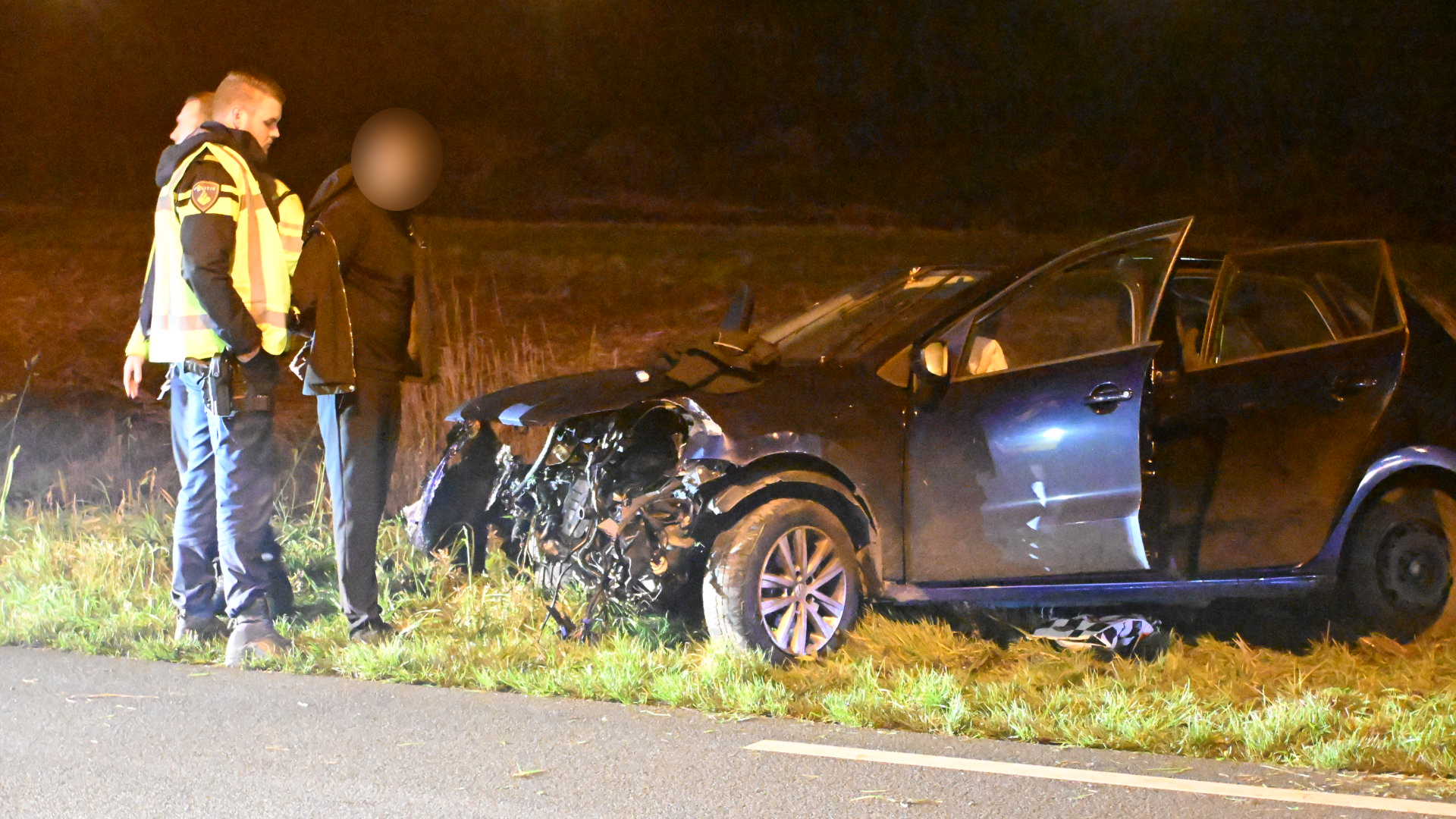 Een auto met ernstige schade aan de voorkant staat langs de weg, terwijl twee politieagenten met een persoon praten.