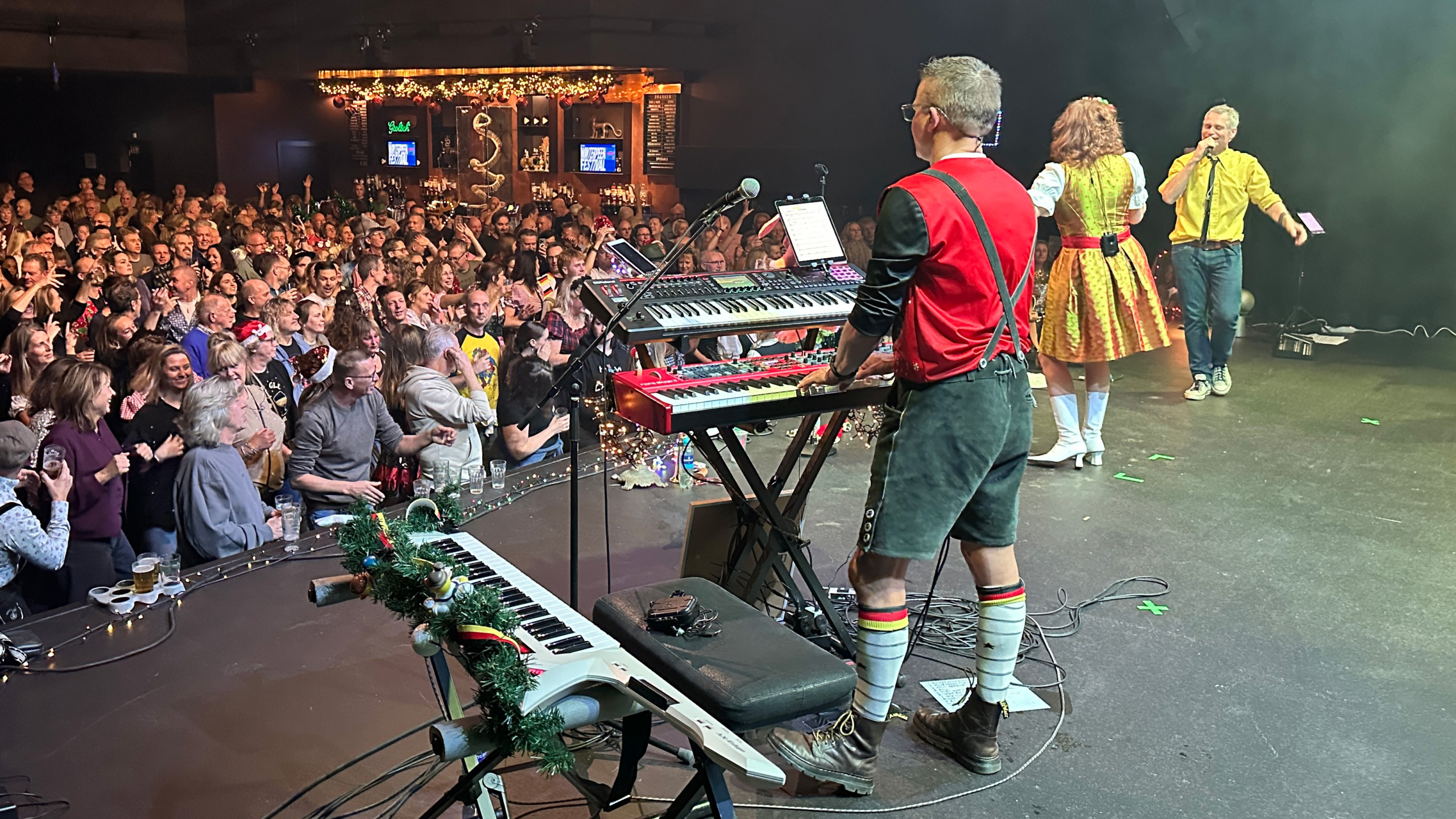 Een muzikant speelt keyboard op een podium voor een enthousiast publiek, met versieringen en andere artiesten.
