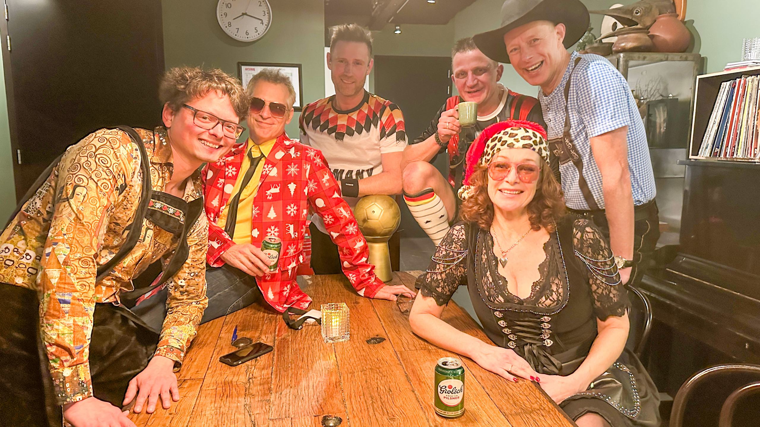 Groep mensen in kleurrijke kleding met feestthema, lachend rond een houten tafel met drankjes en een voetbal.