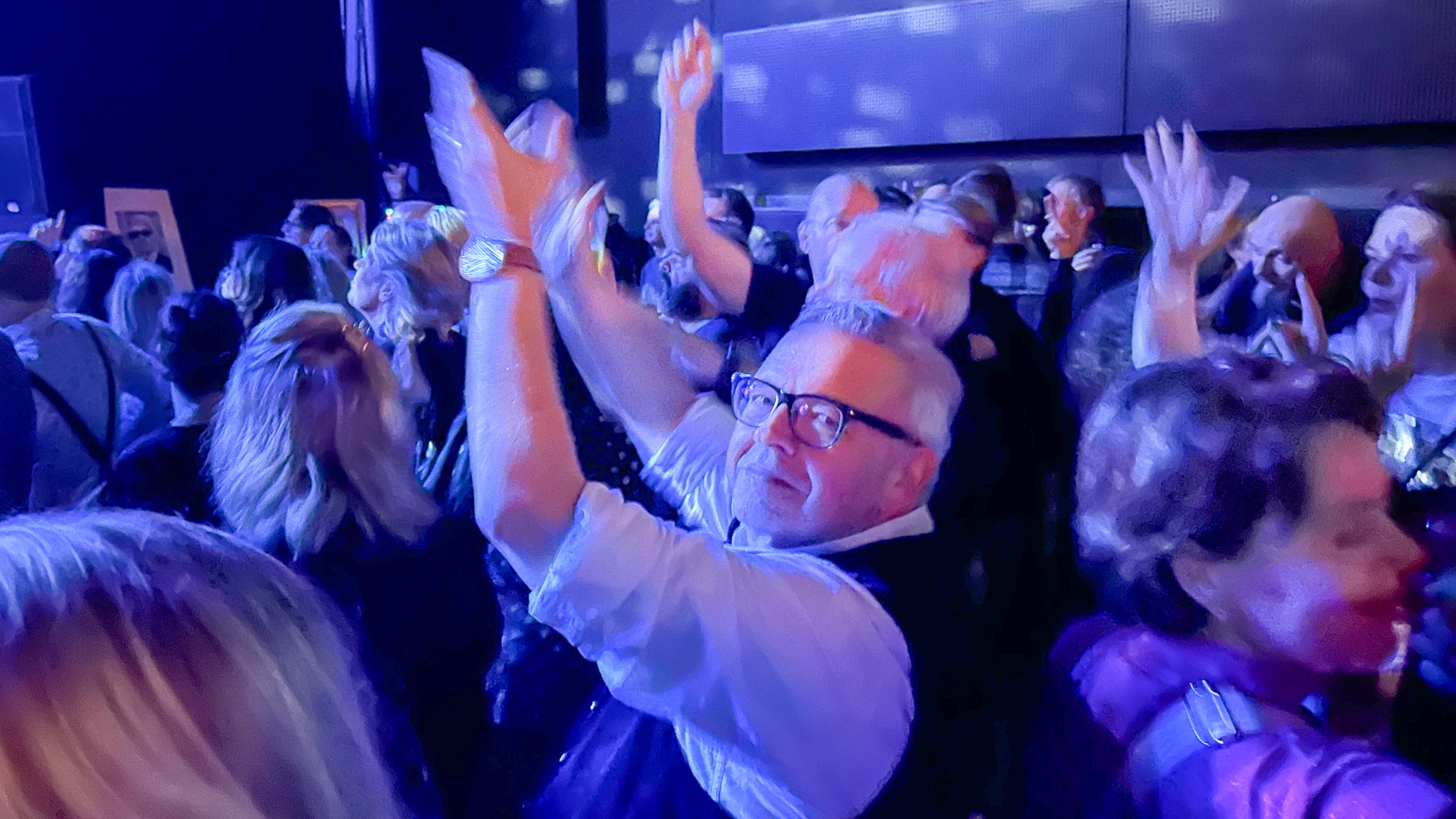 Mensen dansen en applaudisseren bij een feestelijke gelegenheid in een donkere zaal met blauw licht.
