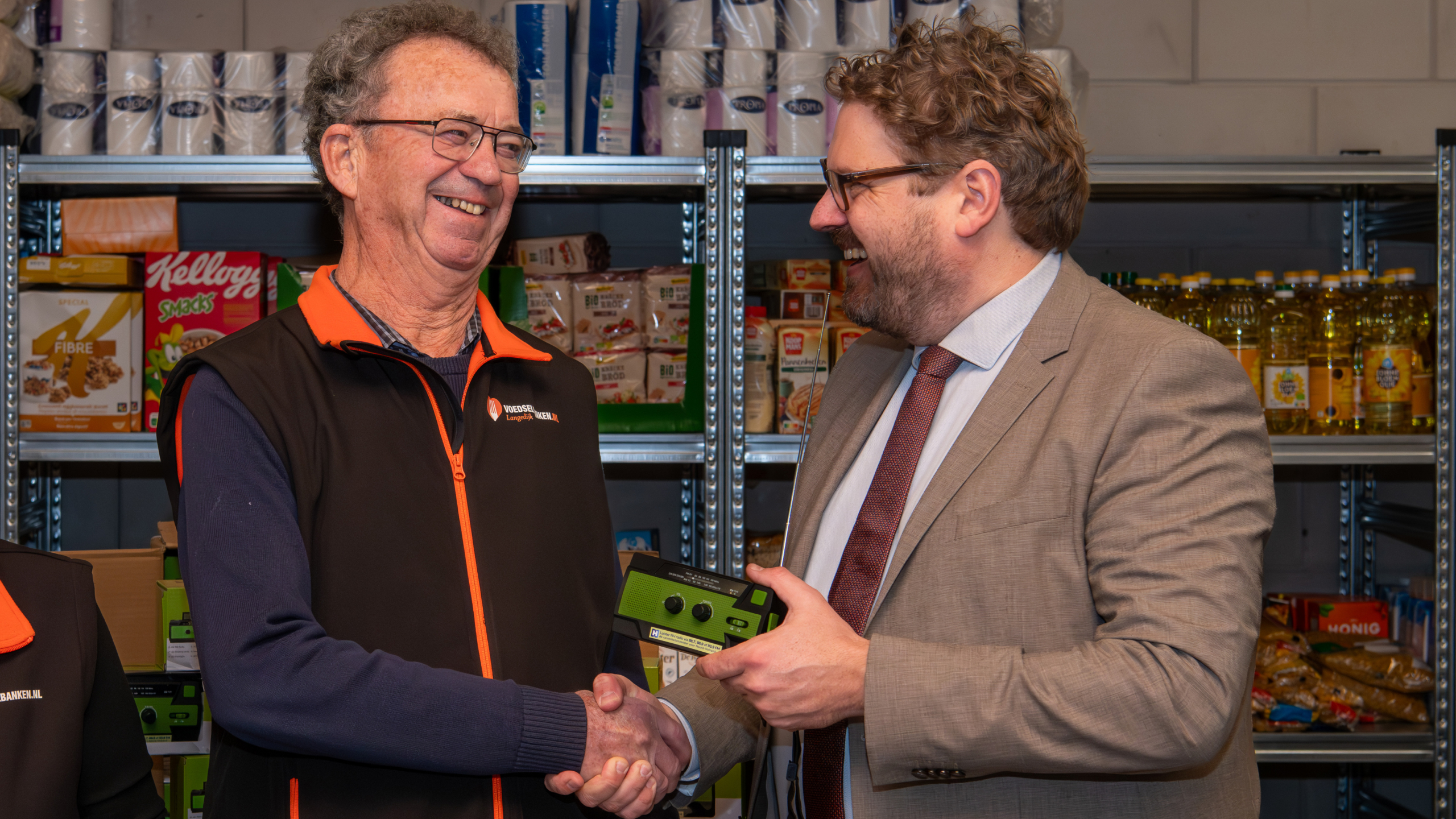 Twee mannen schudden elkaar de hand bij een voedselbank, met op de achtergrond planken gevuld met voedselproducten.