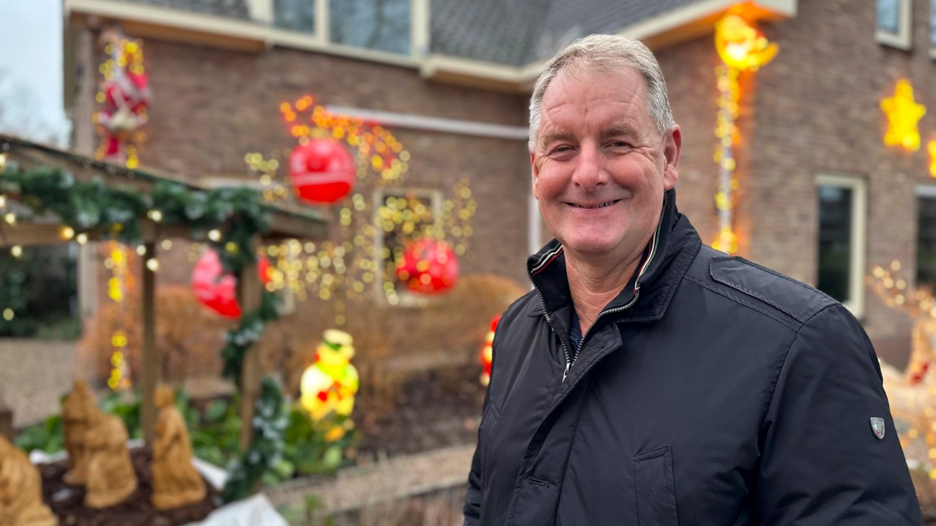 Man in een zwarte jas staat glimlachend voor een huis met kleurrijke kerstdecoraties.
