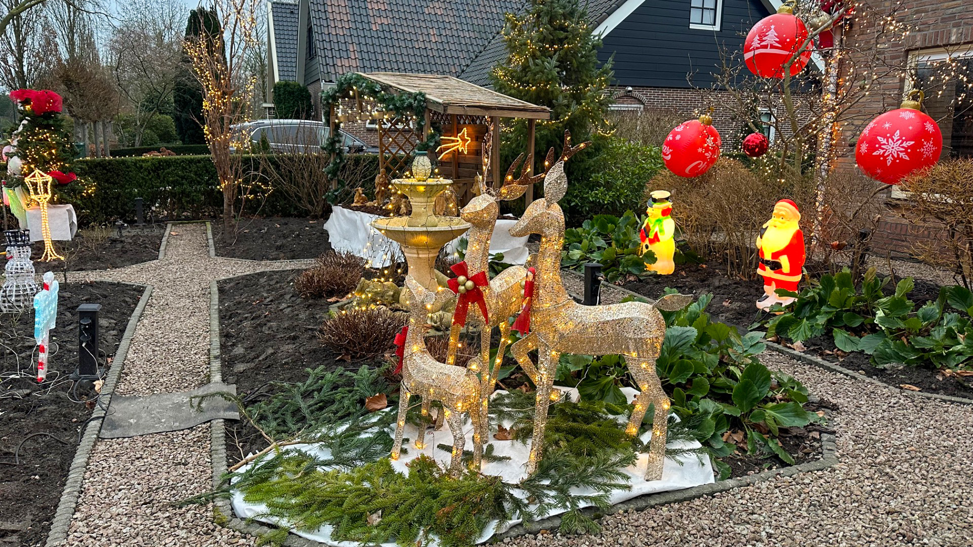 Verlichte kersttuin met rendierfiguren, kerstmanbeelden en rode decoratieballen tussen de groene struiken.
