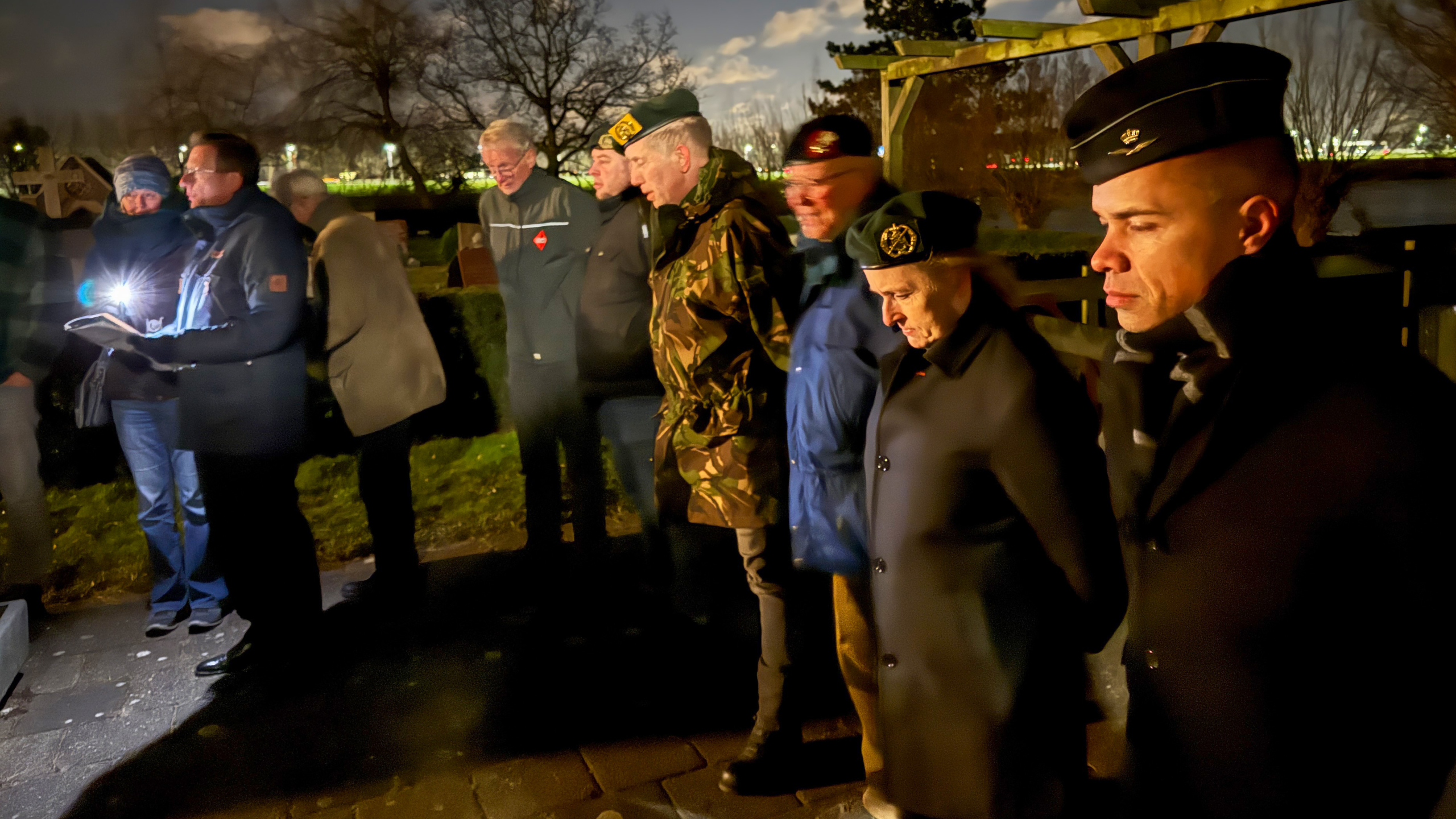 Mensen in uniform staan in een rij tijdens een nachtelijke herdenkingsceremonie met een verlichting in de hand van een persoon.