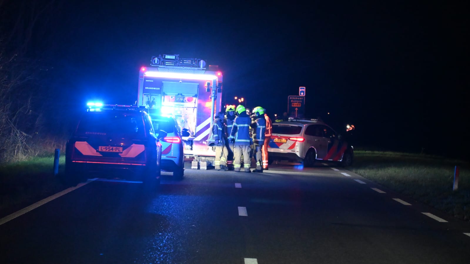Brandweerlieden en politievoertuigen bij nachtelijke noodgeval op een weg met knipperende blauwe lichten.