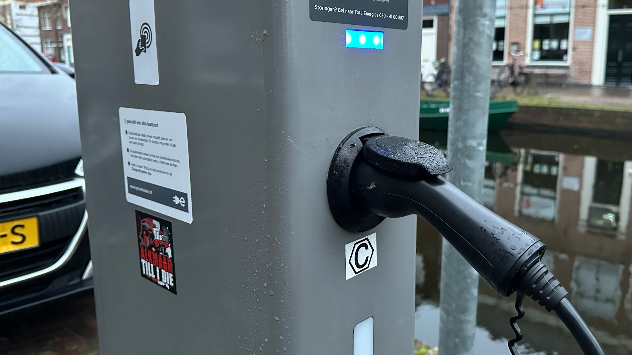 Oplaadpaal voor elektrische voertuigen met een aangesloten stekker, naast een geparkeerde auto en een gracht op de achtergrond.