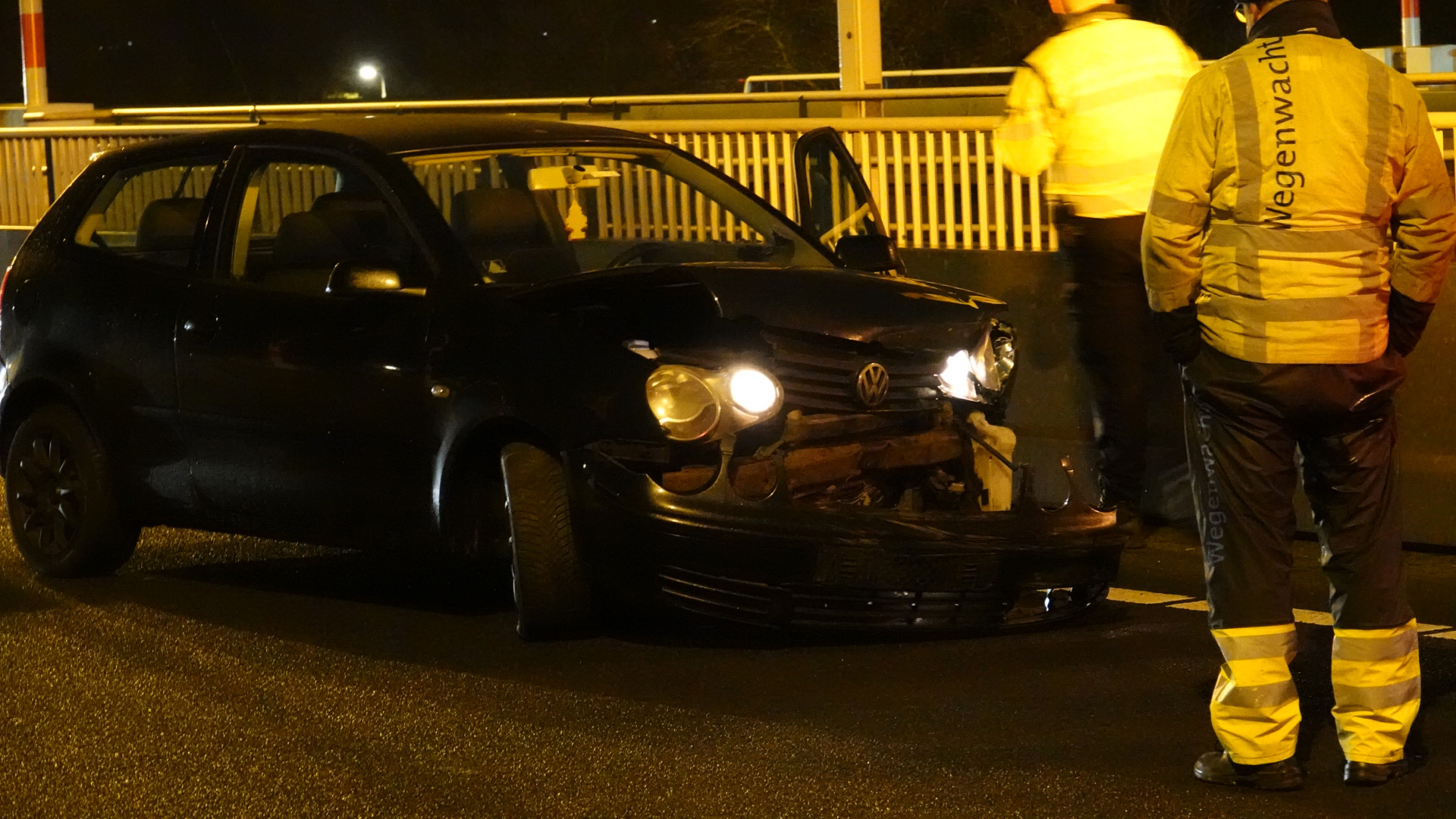 Een beschadigde zwarte auto op een weg met twee wegenwachten die ernaast staan.