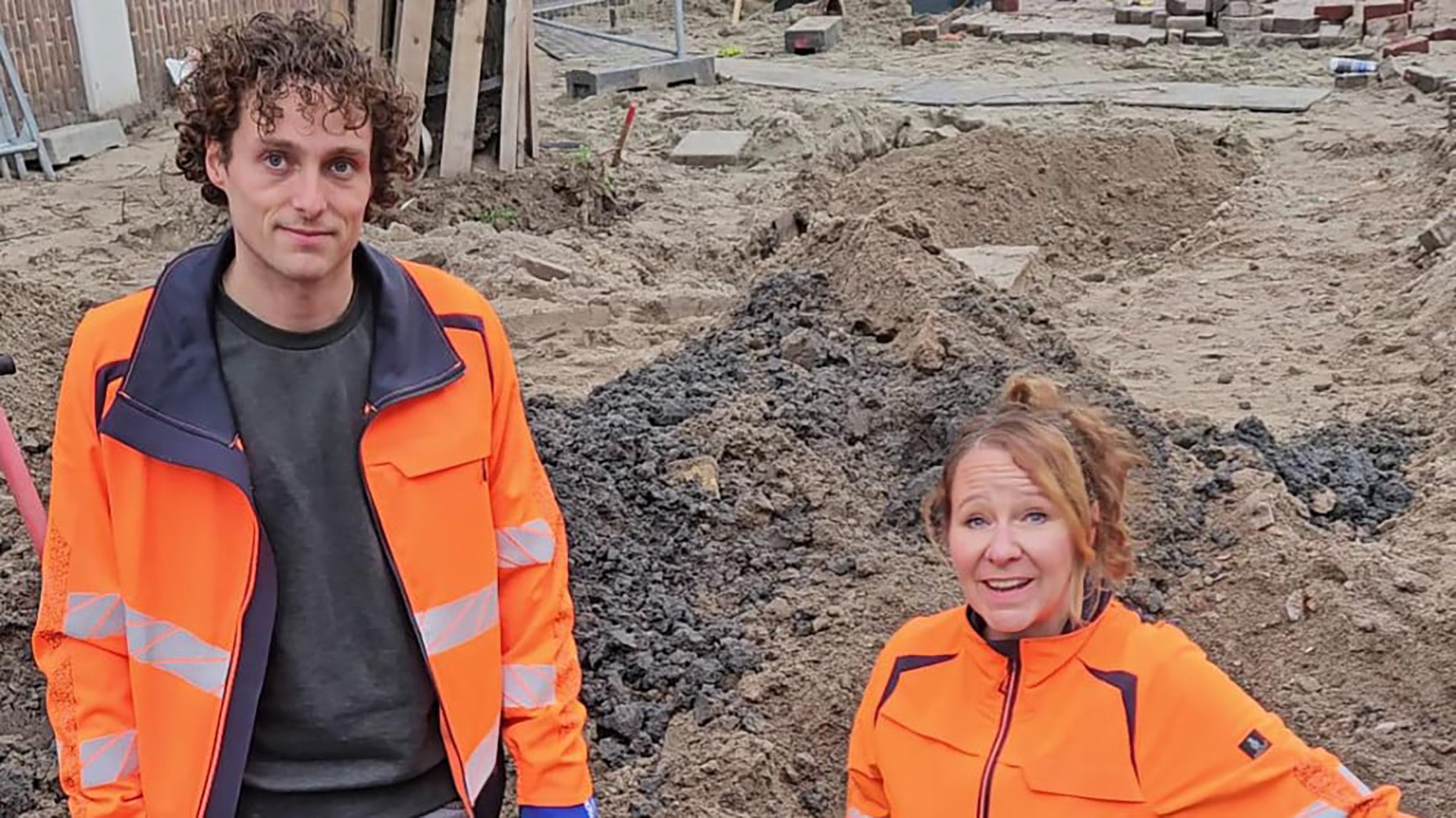Twee mensen in oranje werkkleding op een bouwplaats vol zand en aarde.
