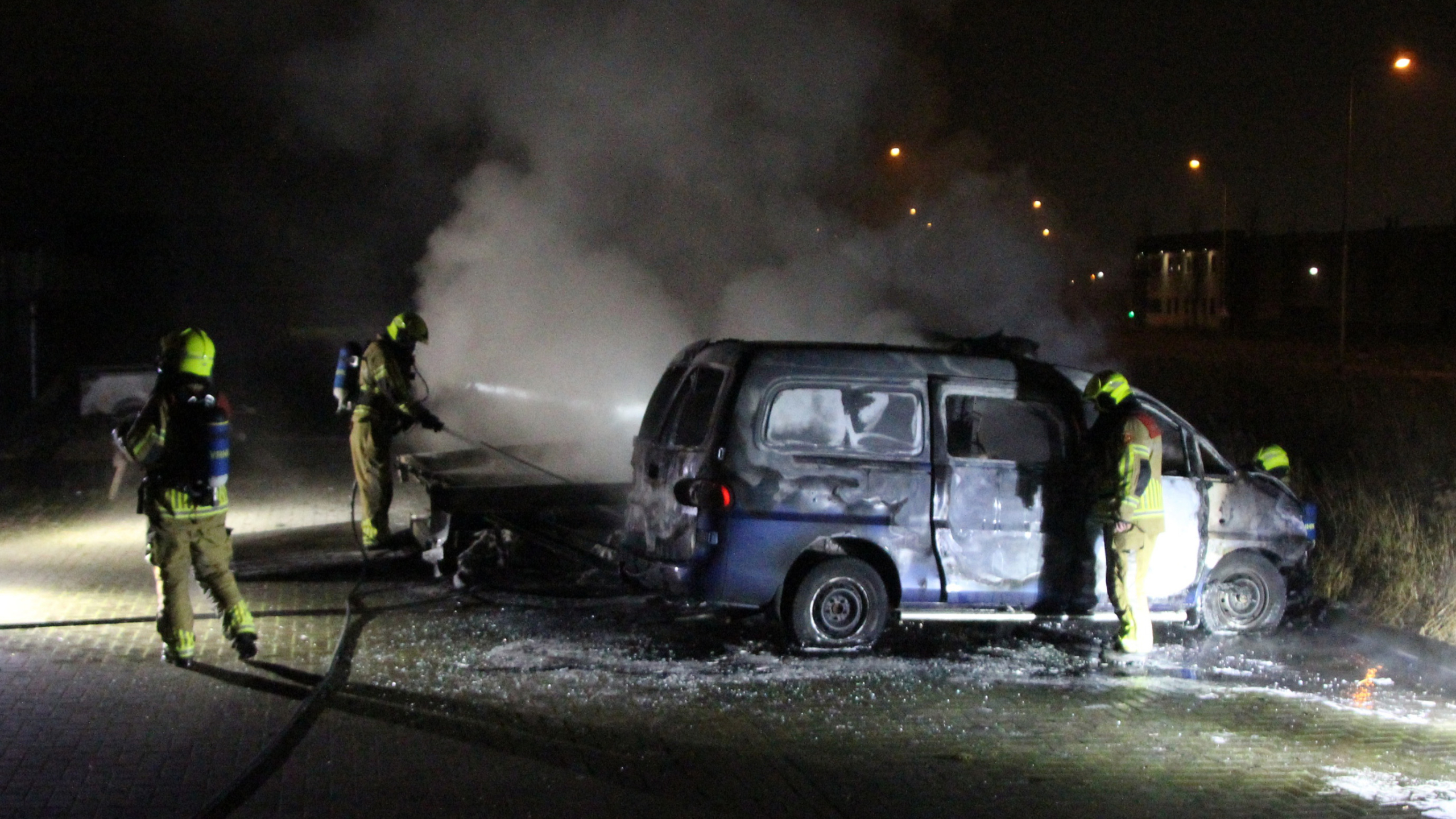 Brandweerlieden blussen een brandende bestelwagen bij nacht, met rook die uit het voertuig opstijgt.