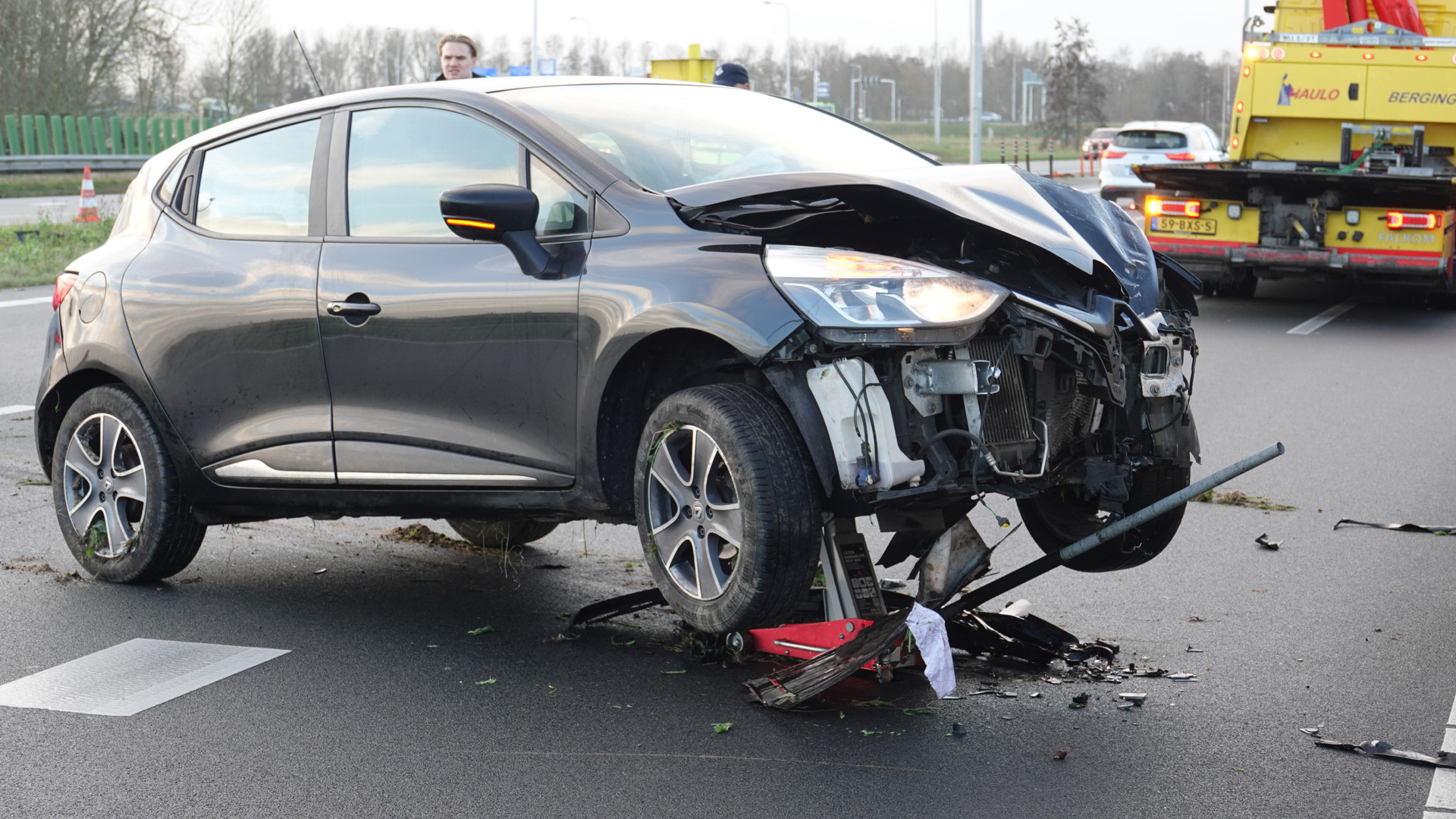Auto met zware schade aan de voorkant na een verkeersongeval, met sleepwagen en omstanders op de achtergrond.
