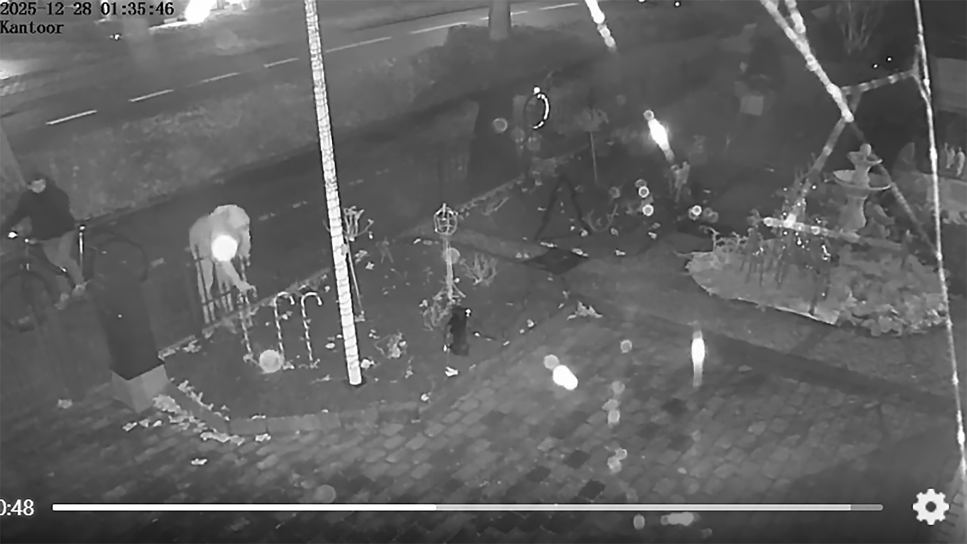 Nachtelijke beveiligingsbeeld van een tuin met decoraties, een persoon op een fiets, en een tijdstempel linksboven.