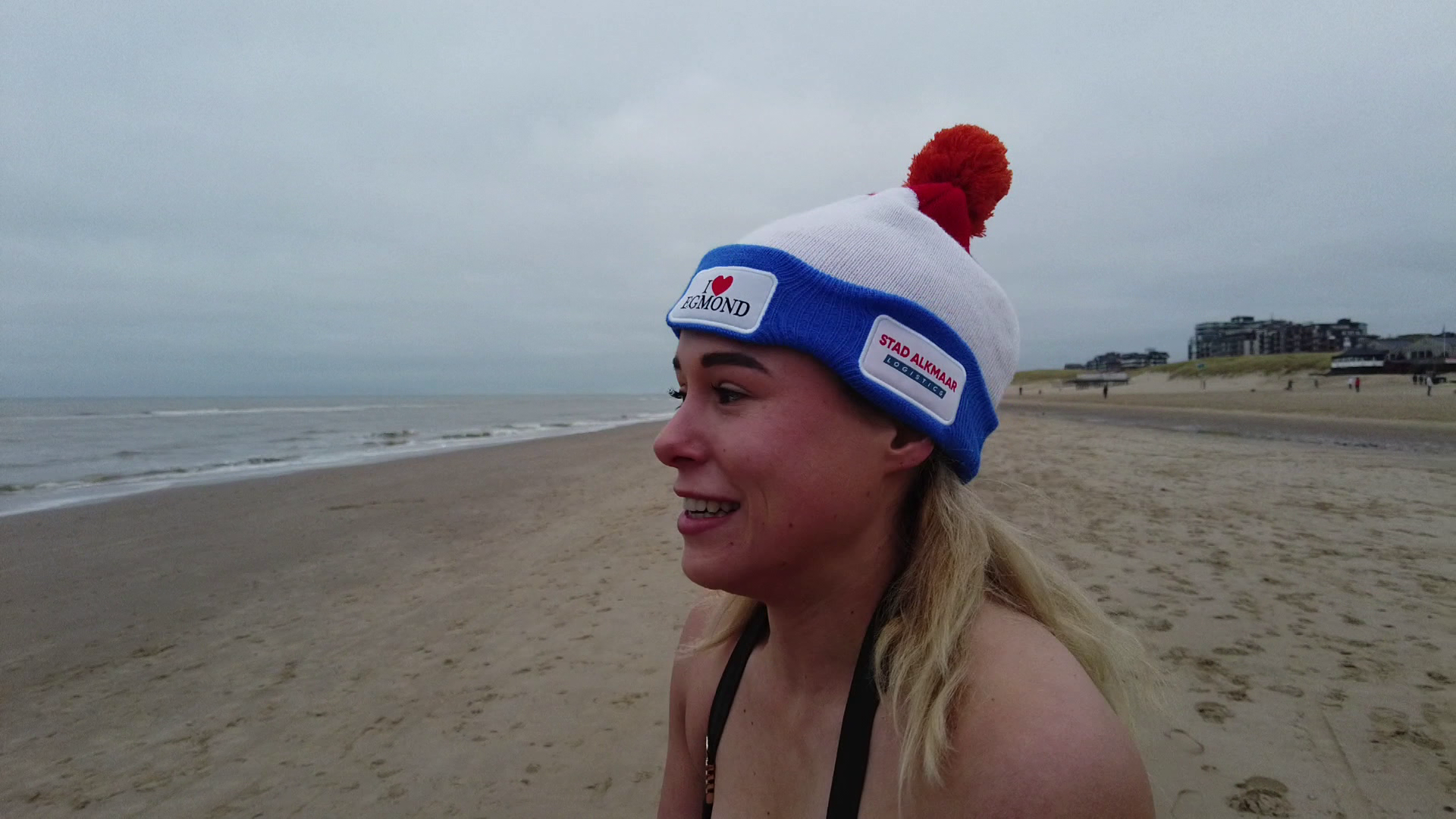 Vrouw aan het strand draagt een witte muts met blauwe rand en rode pompon, met tekst "I love Egmond" en "Stad Alkmaar".