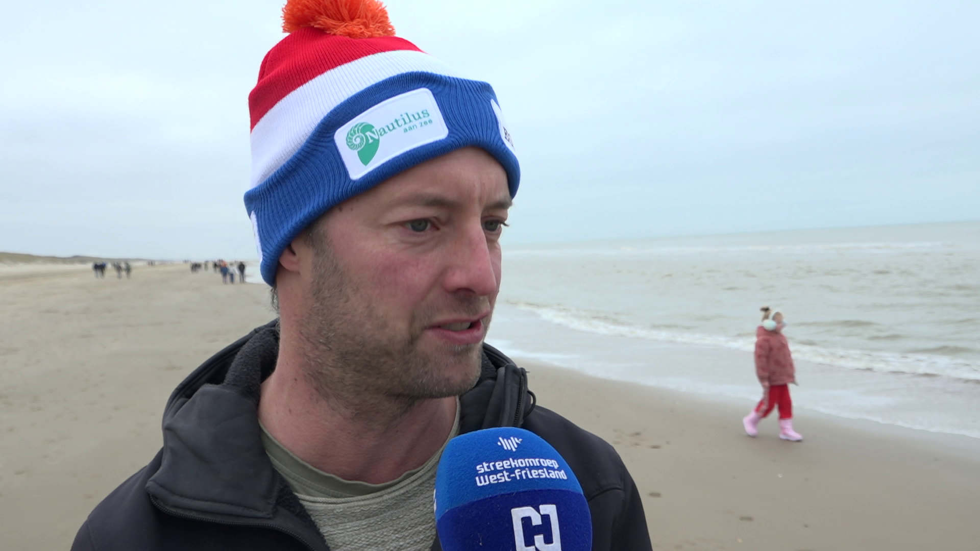 Een man met een kleurrijke muts wordt geïnterviewd op een winterse strandwandeling; een kind loopt achter hem langs.