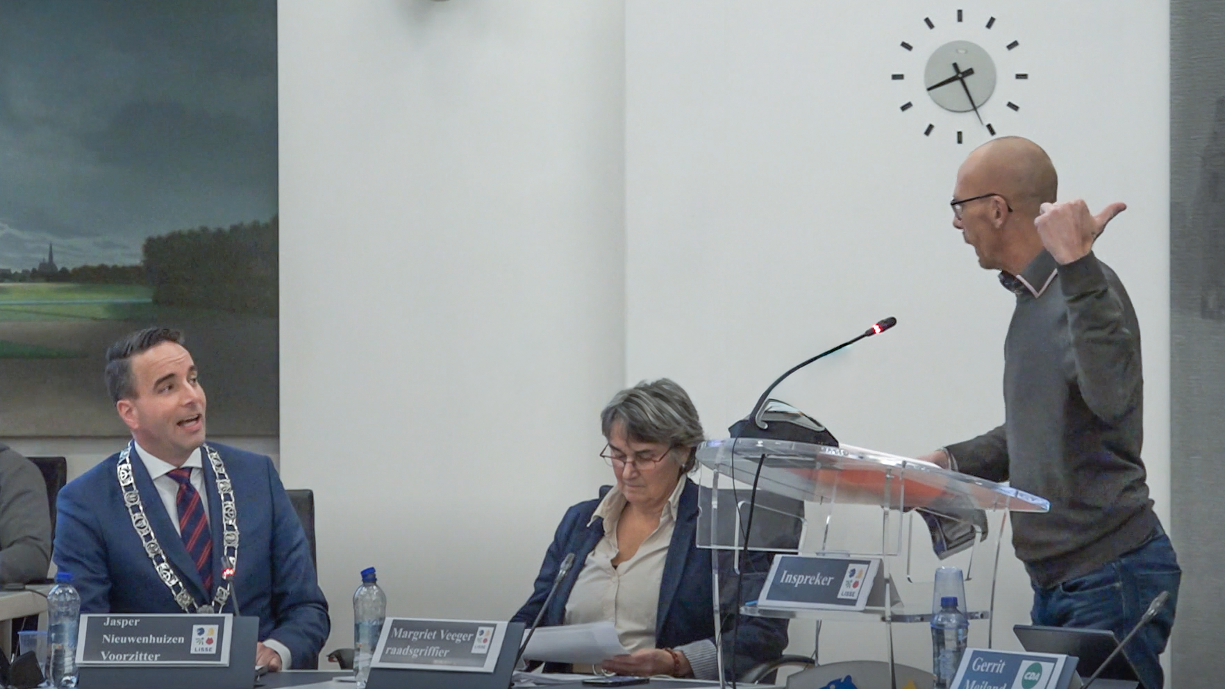 Een man in een grijze trui spreekt gepassioneerd aan een spreekgestoelte in een vergaderzaal, terwijl een man in een blauw pak met een ambtsketen toekijkt en een vrouw achter een tafel bezig is met notities. Op de achtergrond is een schilderij en een klok te zien.