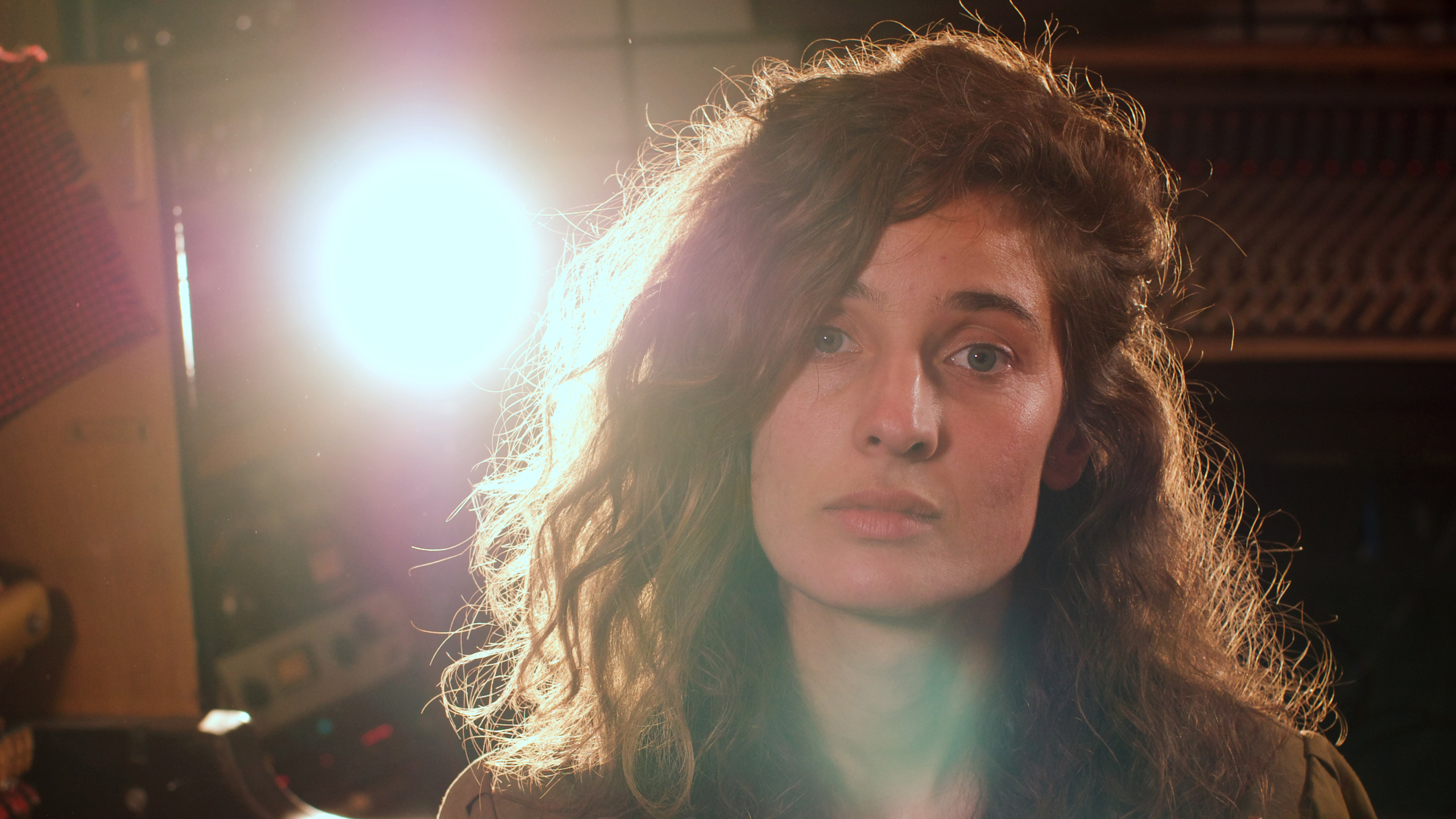 Vrouw met krullend haar kijkt recht in de camera, met fel licht op de achtergrond.