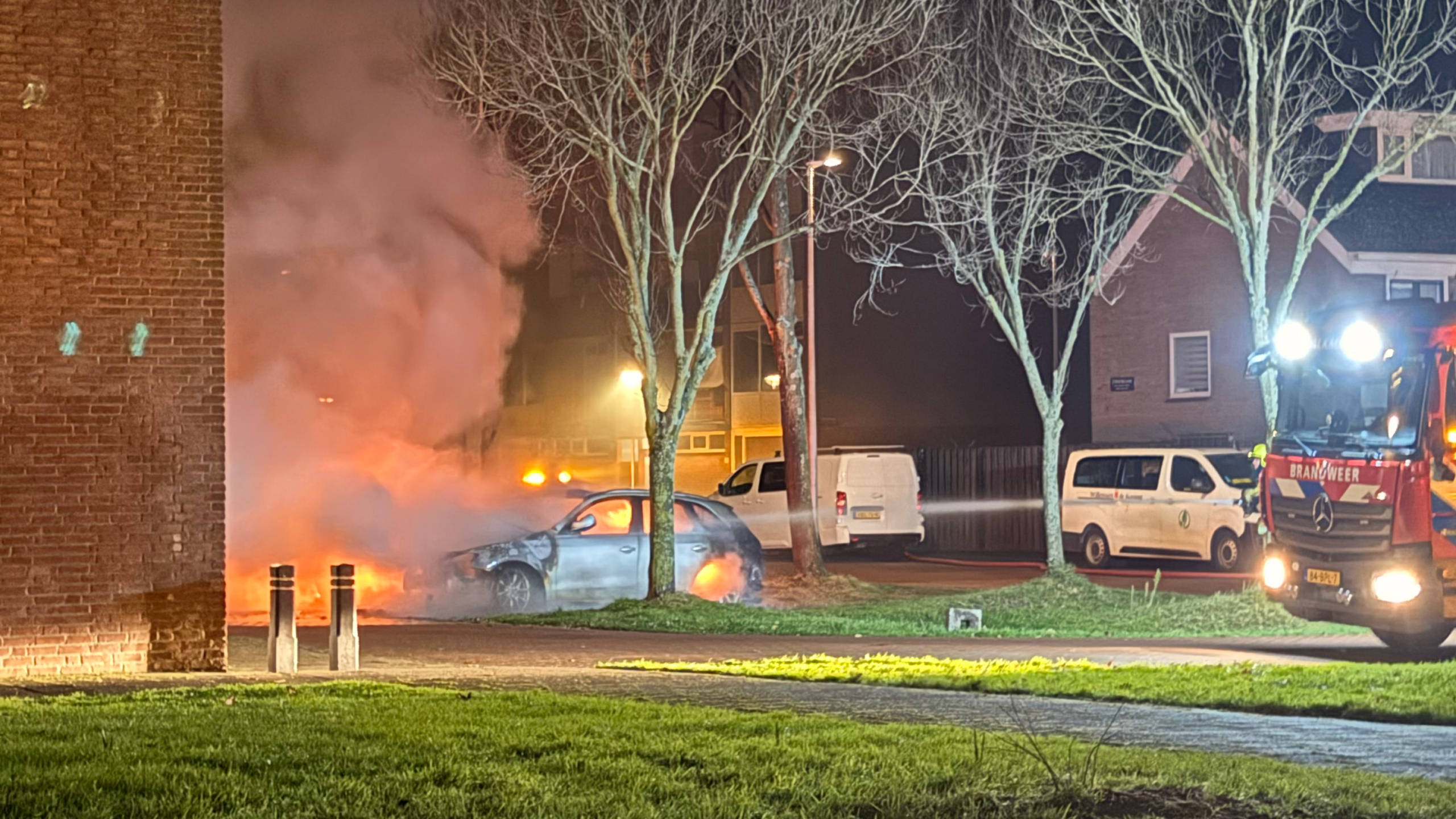 Brandweerwagen en brandende auto omgeven door rook in een woonwijk bij nacht.