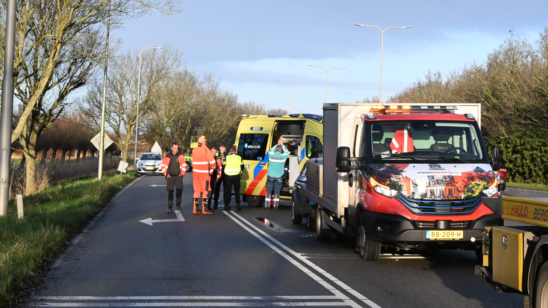 Vrachtwagen en ambulance staan stil op weg met hulpverleners erbij, in een landelijke omgeving met bomen langs de weg.
