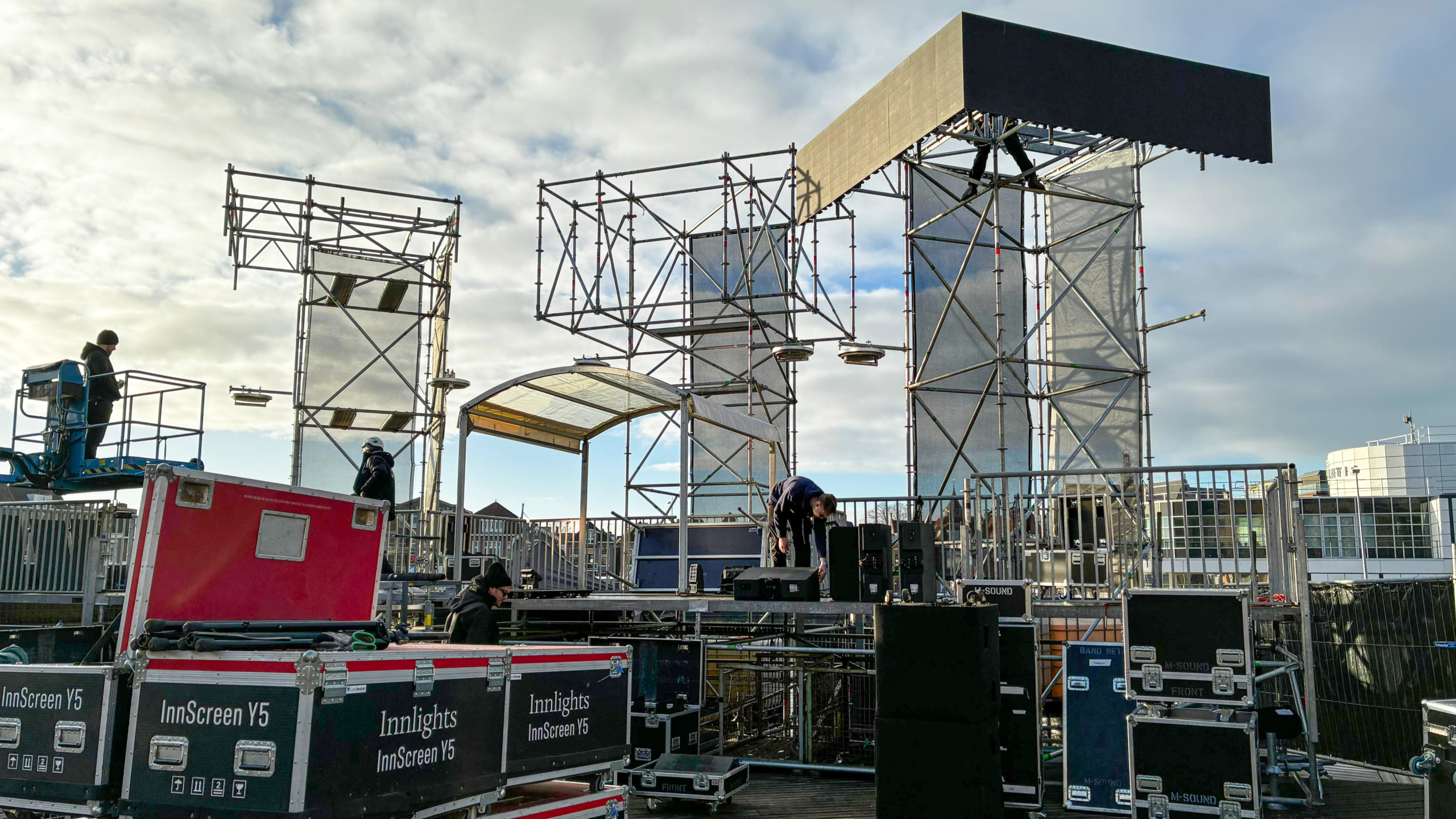 Mensen bouwen een groot podium met metalen steigers en schermen, omringd door apparatuur en apparatuurkisten.