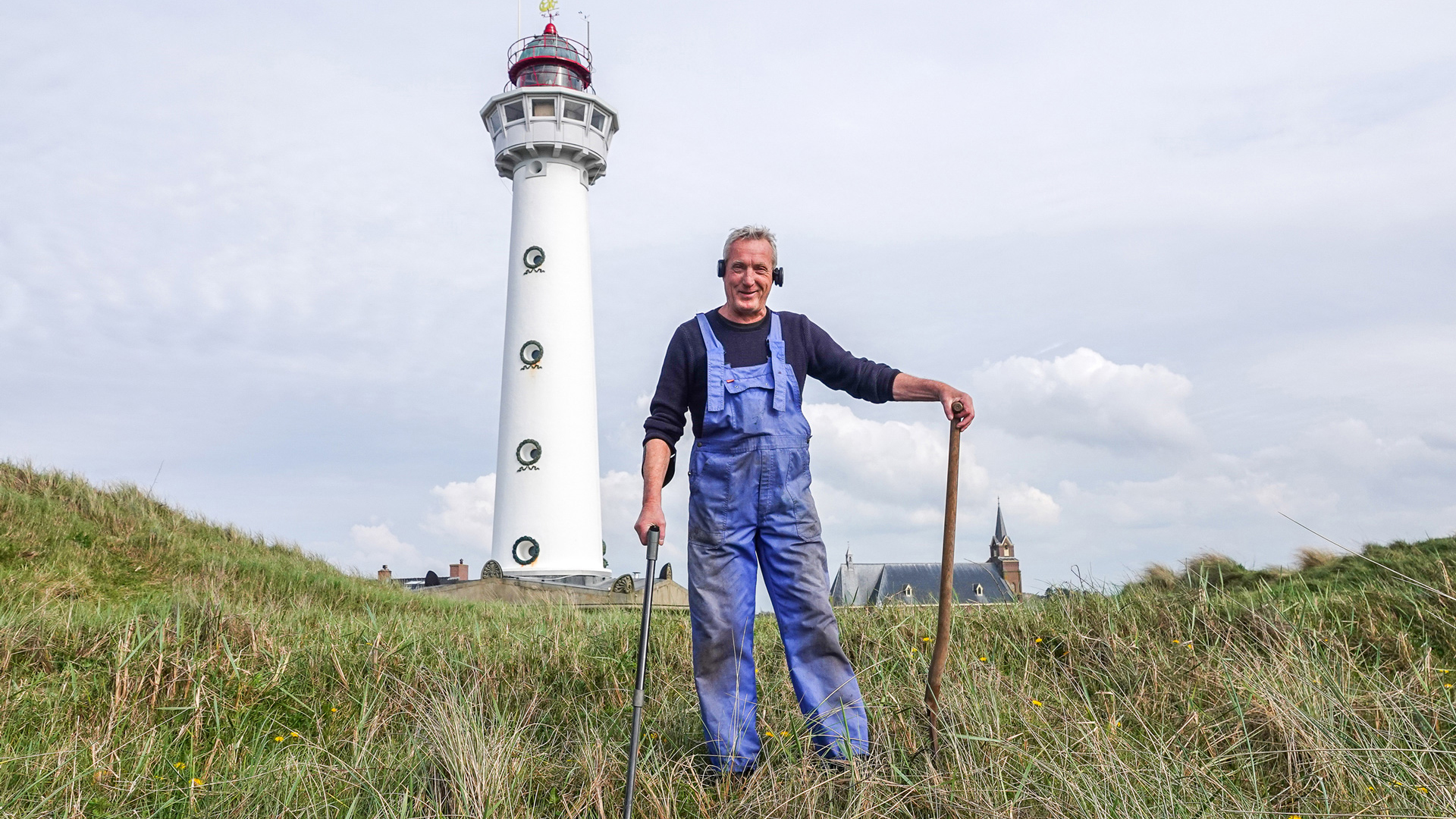 Man in overall met gereedschap, staand op grasveld bij een witte vuurtoren.