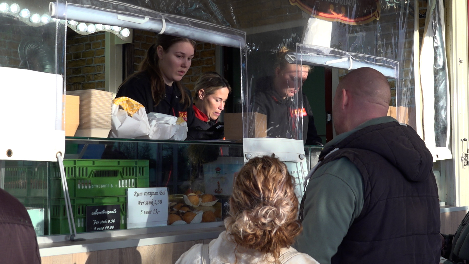 Mensen wachten voor een kraam terwijl verkopers bezig zijn met het inpakken van eten, menuprijzen zichtbaar op de toonbank.