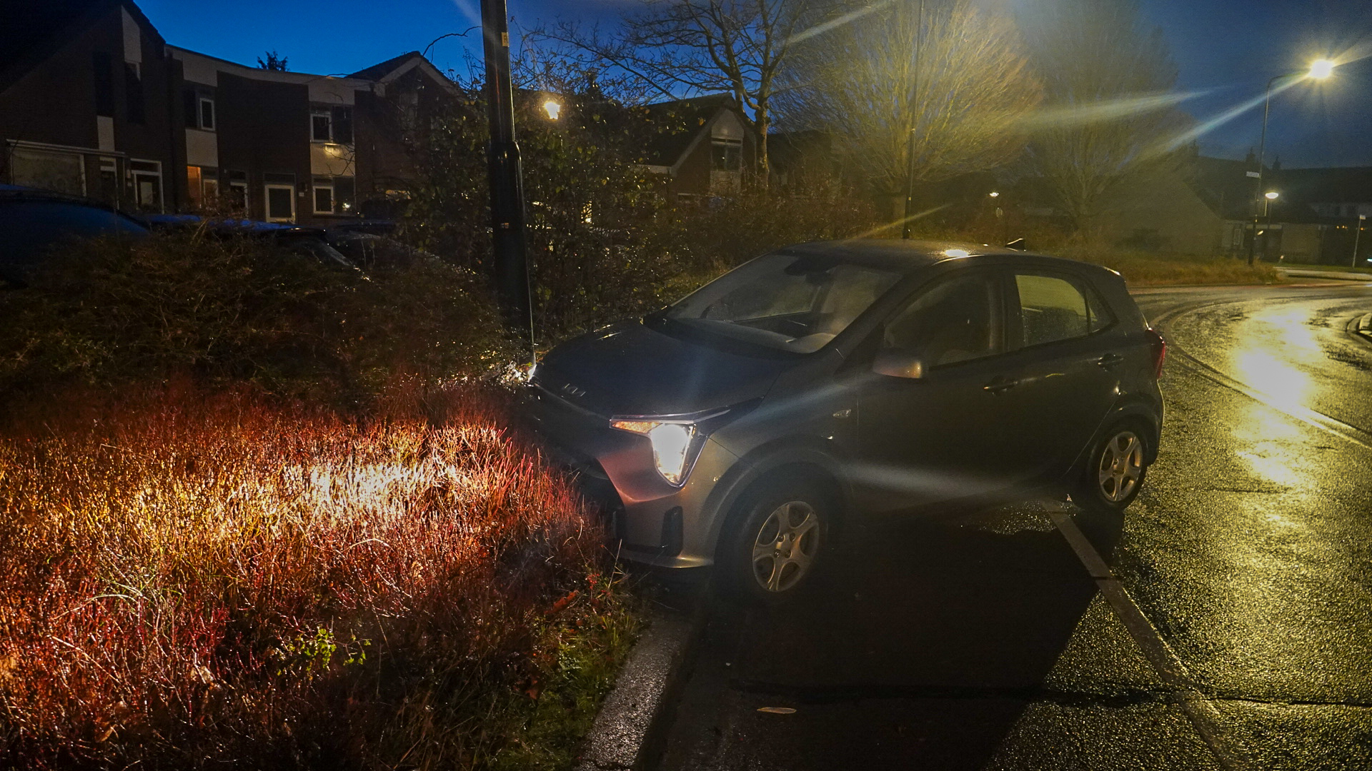 Een auto staat met de voorkant in laag struikgewas langs een natte weg 's avonds met straatverlichting.