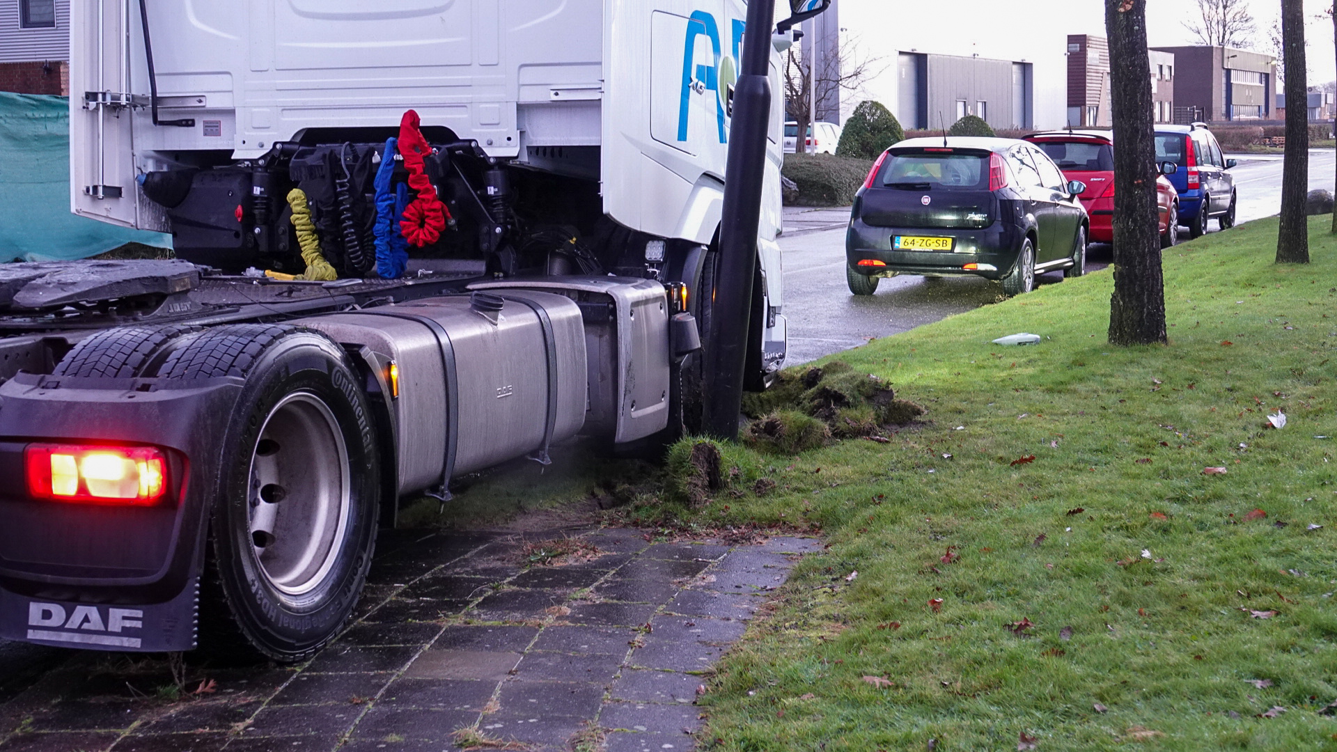 Vrachtwagen is tegen een lantaarnpaal aan gereden op een grasveld naast een stoep, met enkele geparkeerde auto's op de achtergrond.