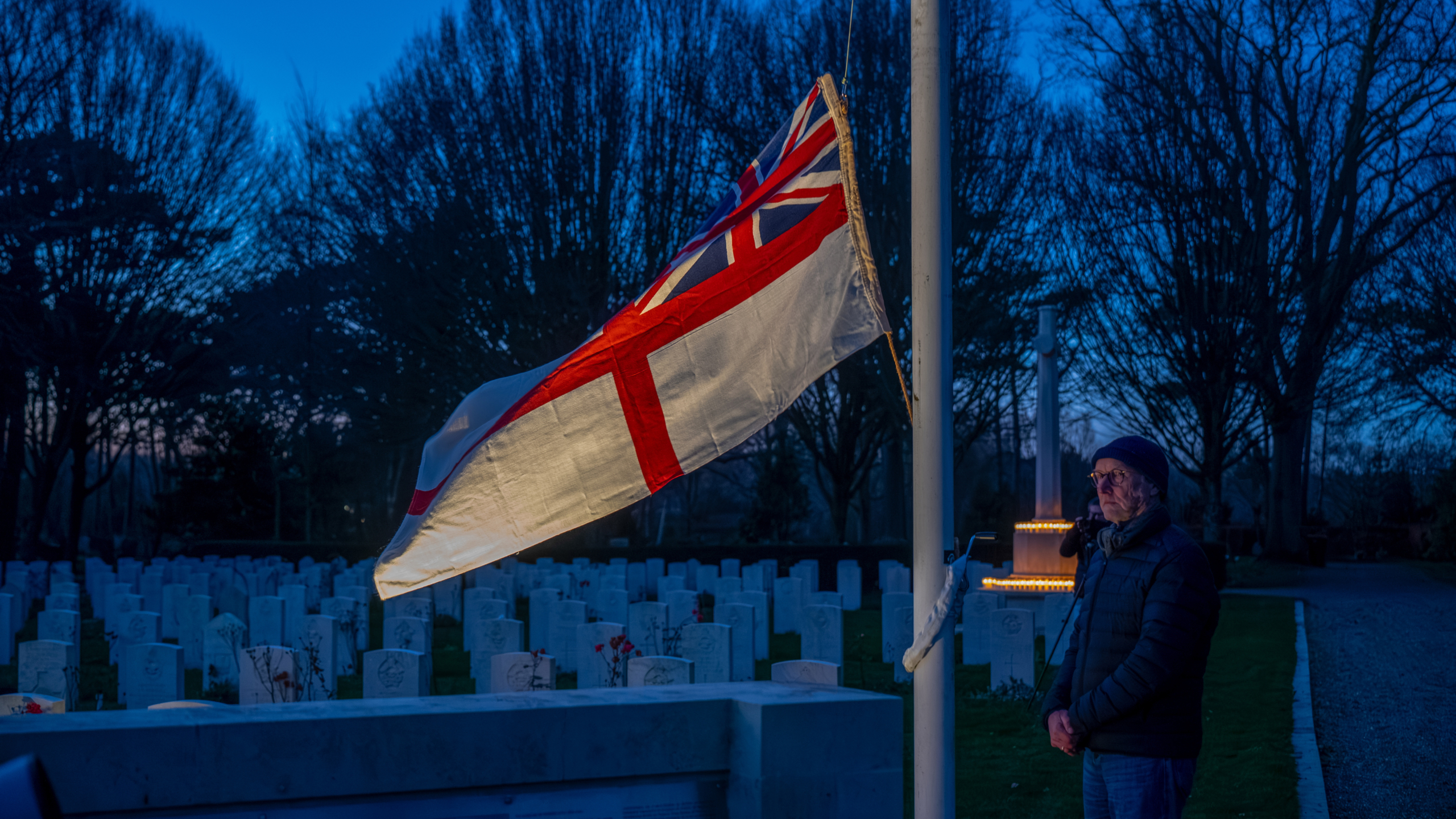 Een oudere man staat in een begraafplaats bij een vlag met een rood kruis en union jack hoek, omgeven door grafstenen en bomen in de schemering.