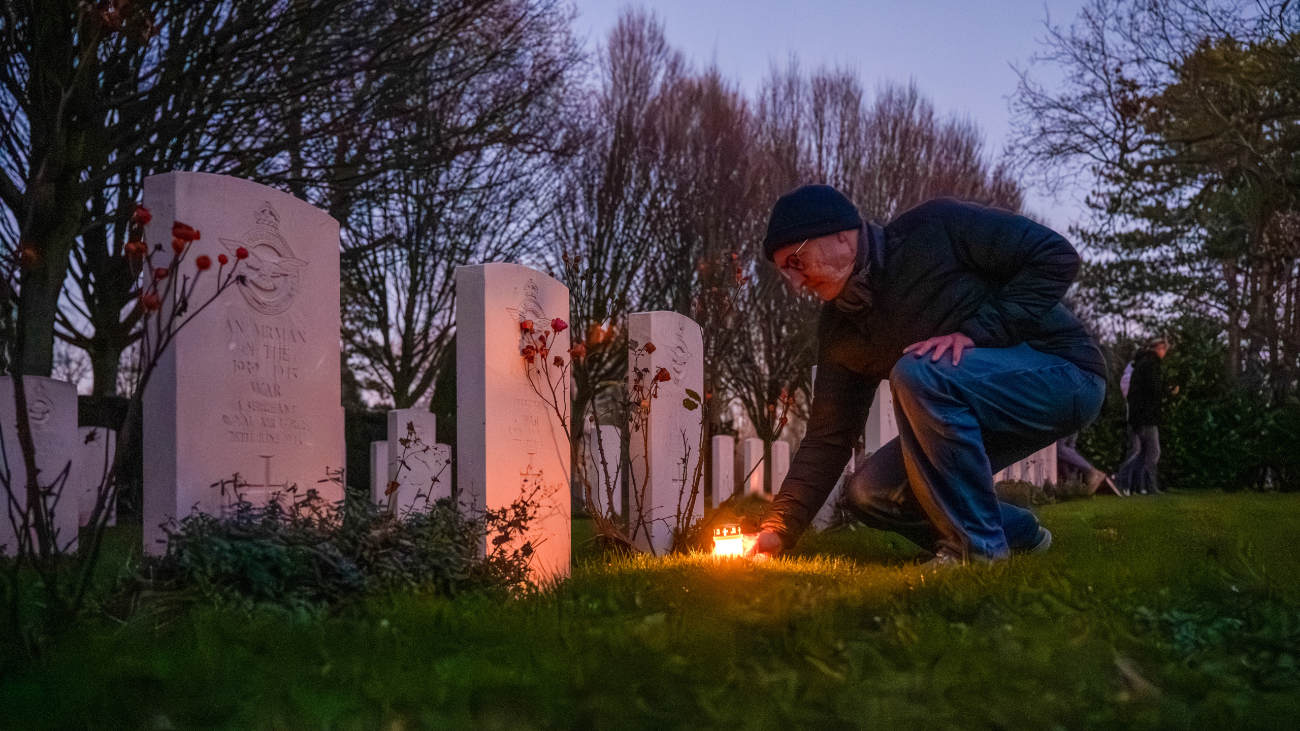 Man steekt een kaars aan bij een militair graf op een schemerige begraafplaats met op de achtergrond bomen en andere bezoekers.