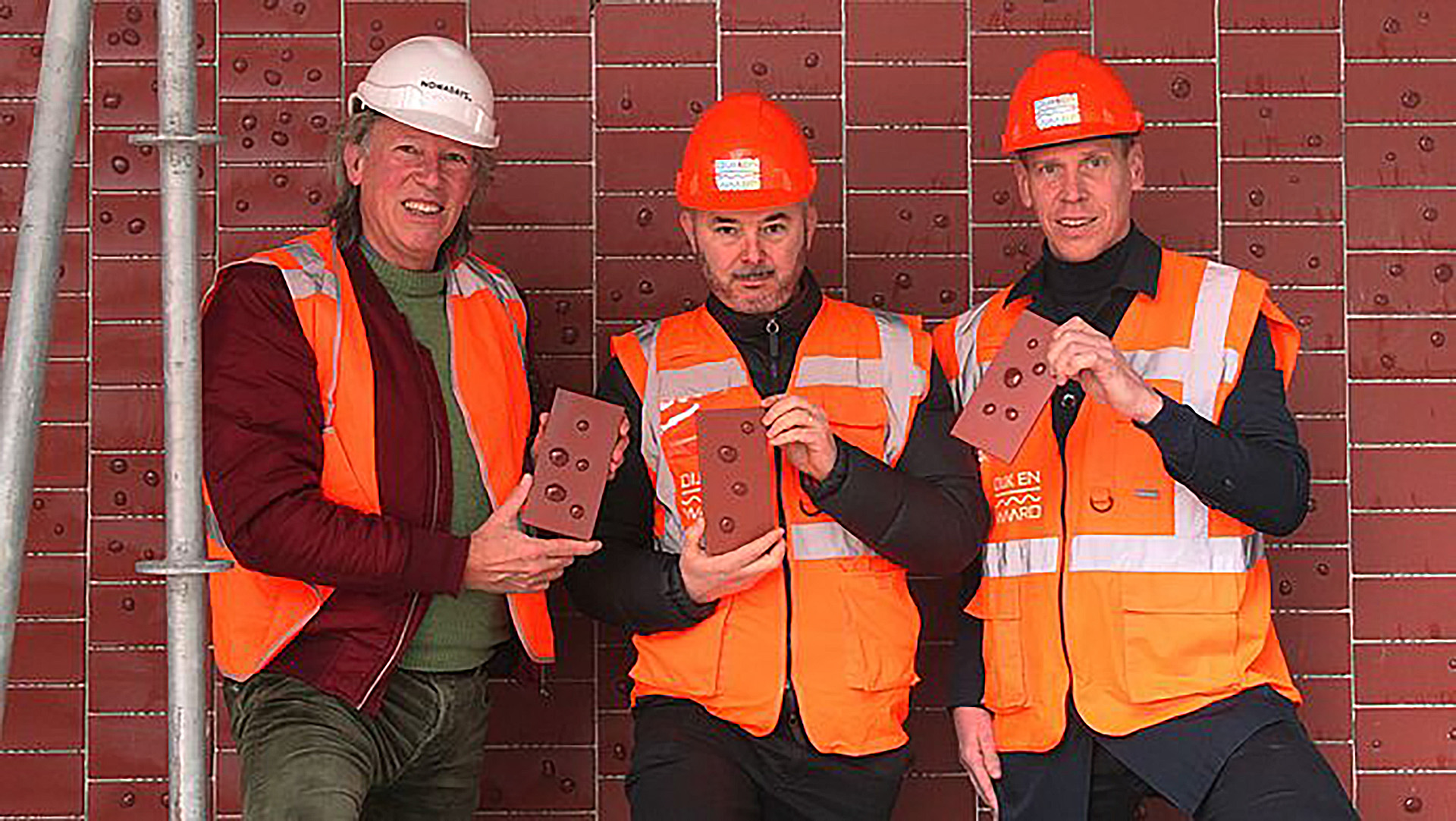 Drie mannen in veiligheidskleding en helmen poseren met bakstenen voor een muur met vergelijkbare stenen.