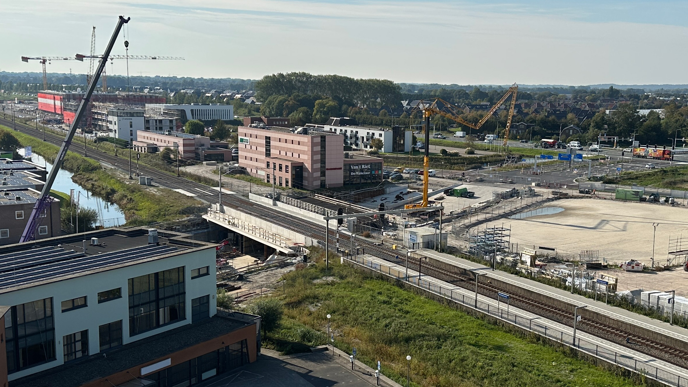 Bouwterrein met hijskranen nabij een spoorlijn en gebouwen onder constructie, omringd door groen en wegen.