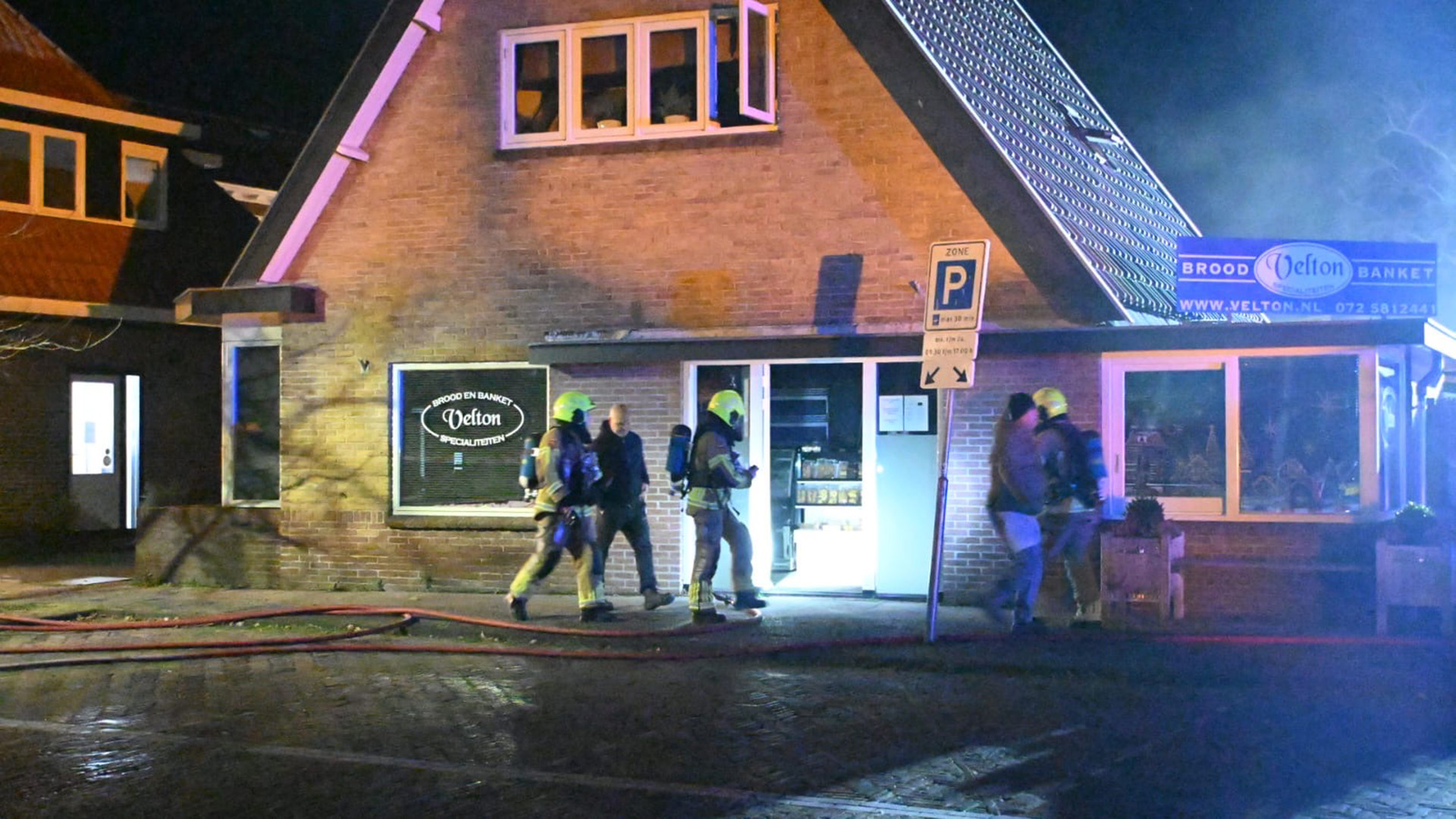 Brandweerlieden bij een bakkerij met rookslangen op de stoep, 's avonds verlicht door straatverlichting.