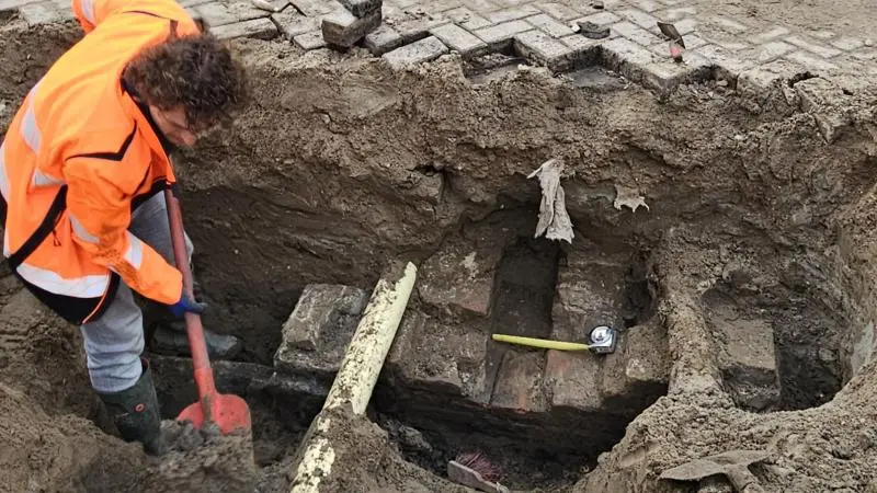 Een persoon in een oranje veiligheidsvest graaft met een schep in een bouwput met bakstenen en gereedschap.